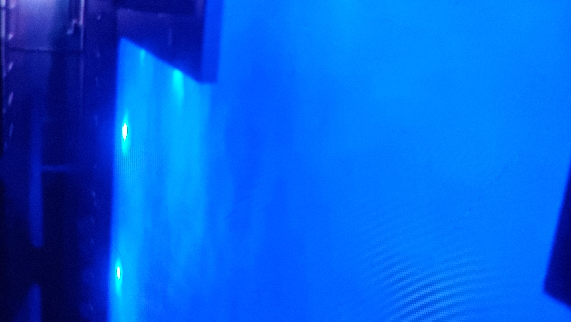 The image depicts a dark environment illuminated by bright blue lights. Several circular light sources are visible at the far end of the frame, casting pools of light onto a dark, reflective surface. The overall impression is of an underwater scene or a dimly lit space with artificial lighting. The location is not specified in the image.