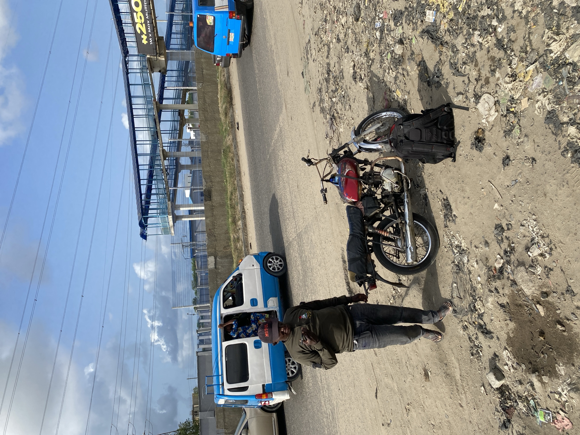 The image shows a street scene in Lekki, Nigeria. A man wearing a black cap, green long-sleeved shirt, and jeans stands next to a motorcycle lying on its side amidst scattered trash along the edge of the road. Behind him is a white and blue van parked on the road. In the background, there's an elevated structure with billboards and overhead power lines against a partly cloudy sky. Another blue vehicle is visible in the distance. The ground is mostly dirt and debris.