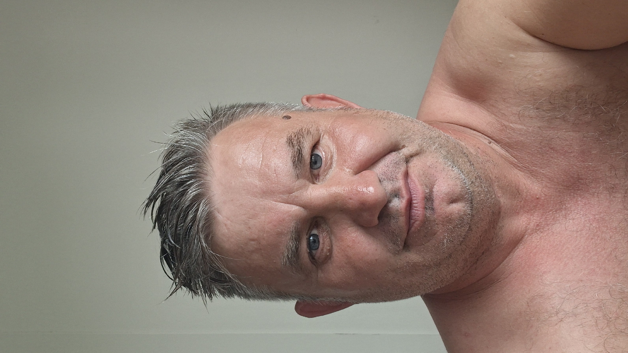 The image is a close-up, low-angle selfie of a middle-aged man with graying hair and blue eyes. He is looking directly at the camera with a slight smile. The man is shirtless, and his skin appears flushed, possibly from heat or exertion. There is a mole on his forehead. The background is a plain, light-colored wall, suggesting an indoor setting. The lighting is even and appears to be from an overhead source, indicating it is likely daytime. No text is visible in the image. The orientation of the image is sideways, with the man's head to the right.