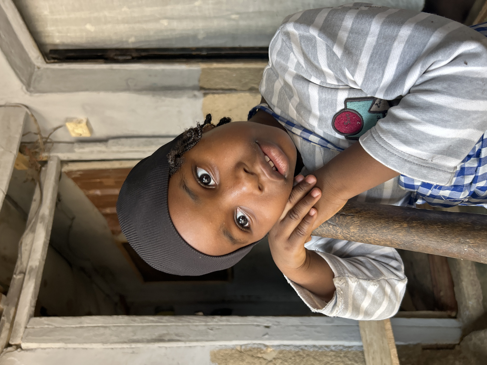 A young person with dark skin and a black head covering is seen from a low angle. They have dark eyes and rest their face on their hands, which are placed on a weathered, wooden railing. The person wears a striped gray and white shirt with a small patch on the chest. The background includes a mix of worn building materials and a window frame. The location is Jos, Nigeria.