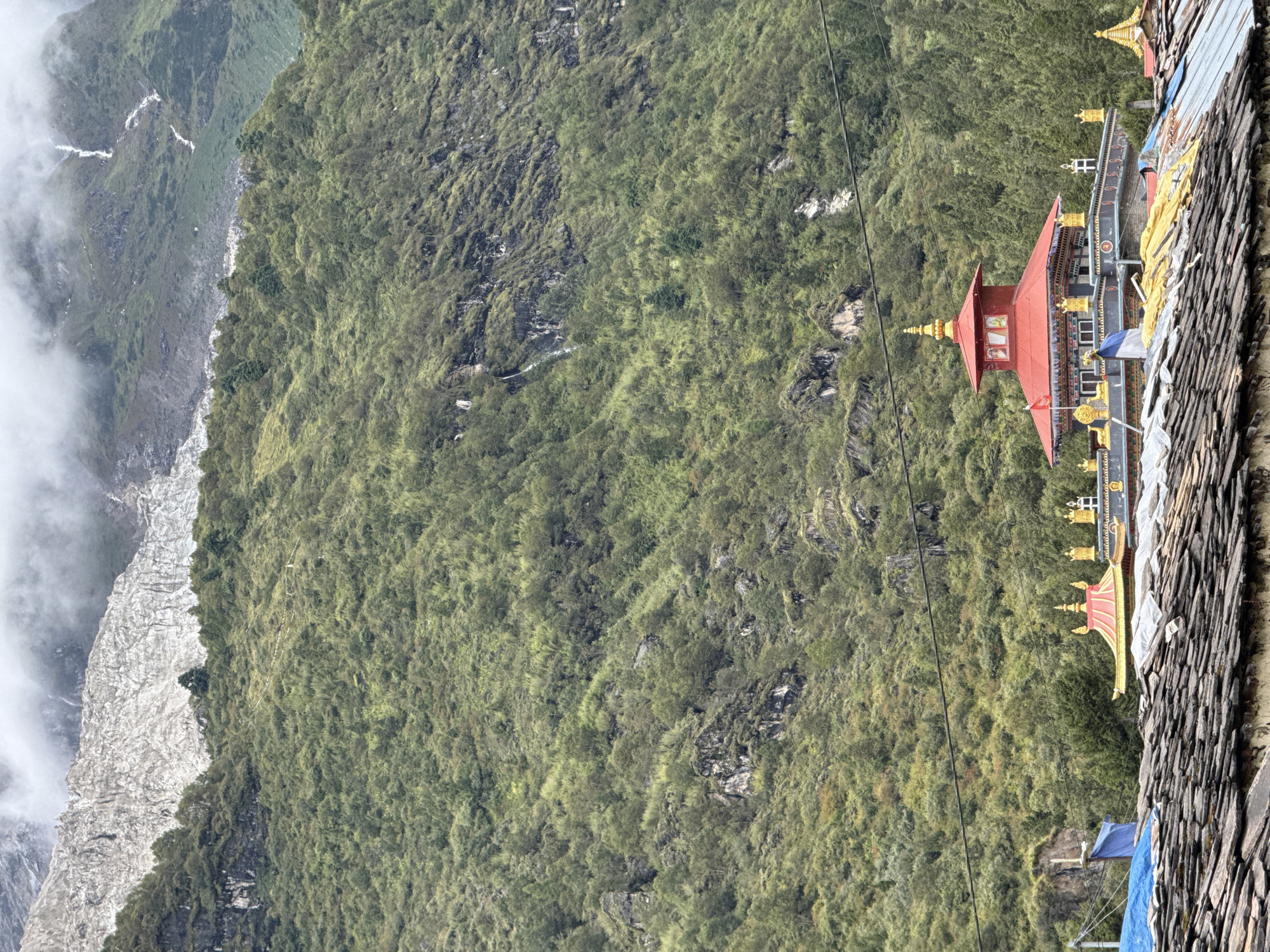The image shows a view of the town of Samagaon, Nepal, with the backdrop of the Himalayas. In the foreground, there are several traditional Tibetan-style buildings with ornate golden roofs and red accents. The buildings are clustered together, and some have prayer flags flying from them. The roofs are made of slate tiles. In the middle ground, a lush green, forested mountainside rises steeply. A narrow, winding path with white markers can be seen traversing the slope. In the background, a massive, jagged glacier dominates the scene, its white ice contrasting with the grey, rocky peaks. Wisps of clouds obscure the top of the glacier and the surrounding mountains. A thin power line stretches across the middle of the image.