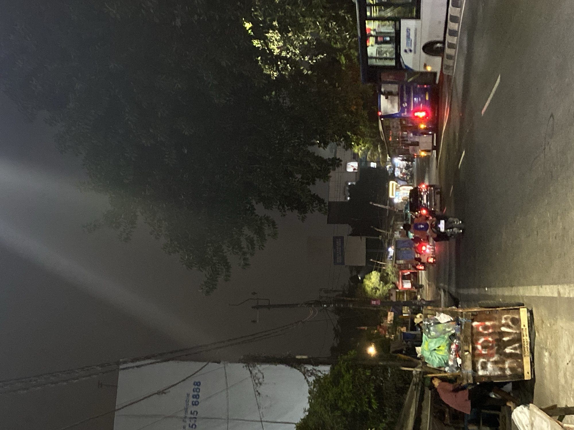 The image depicts a street scene at night in Tangerang, Indonesia. A dimly lit street is lined with trees and buildings. A cart filled with refuse is present on the left side of the road, with a person attending to it. Vehicles, including motorcycles and buses, are on the road, some with their headlights and taillights illuminated. The overall atmosphere is that of a busy urban environment at night.