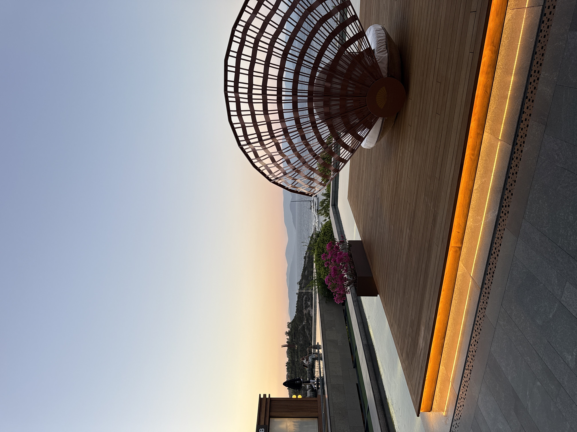 The image captures a serene twilight scene at a waterfront location in Bodrum, Turkey. The foreground features a modern wooden deck with embedded orange lighting, adjacent to a reflective pool of water. A large, fan-shaped rattan structure, serving as a unique seating area with a white cushion, is positioned on the deck. In the midground, a low building with a wooden facade and a black umbrella suggests a cafe or restaurant. Beyond the pool, a scenic view unfolds with rolling hills, a body of water, and distant mountains, all bathed in the soft hues of the setting sun. Several boats are visible on the water, indicating a coastal setting. The sky transitions from a pale blue at the top to warm orange and pink tones near the horizon. The overall atmosphere is tranquil and upscale, typical of a luxury resort or dining establishment in Bodrum.