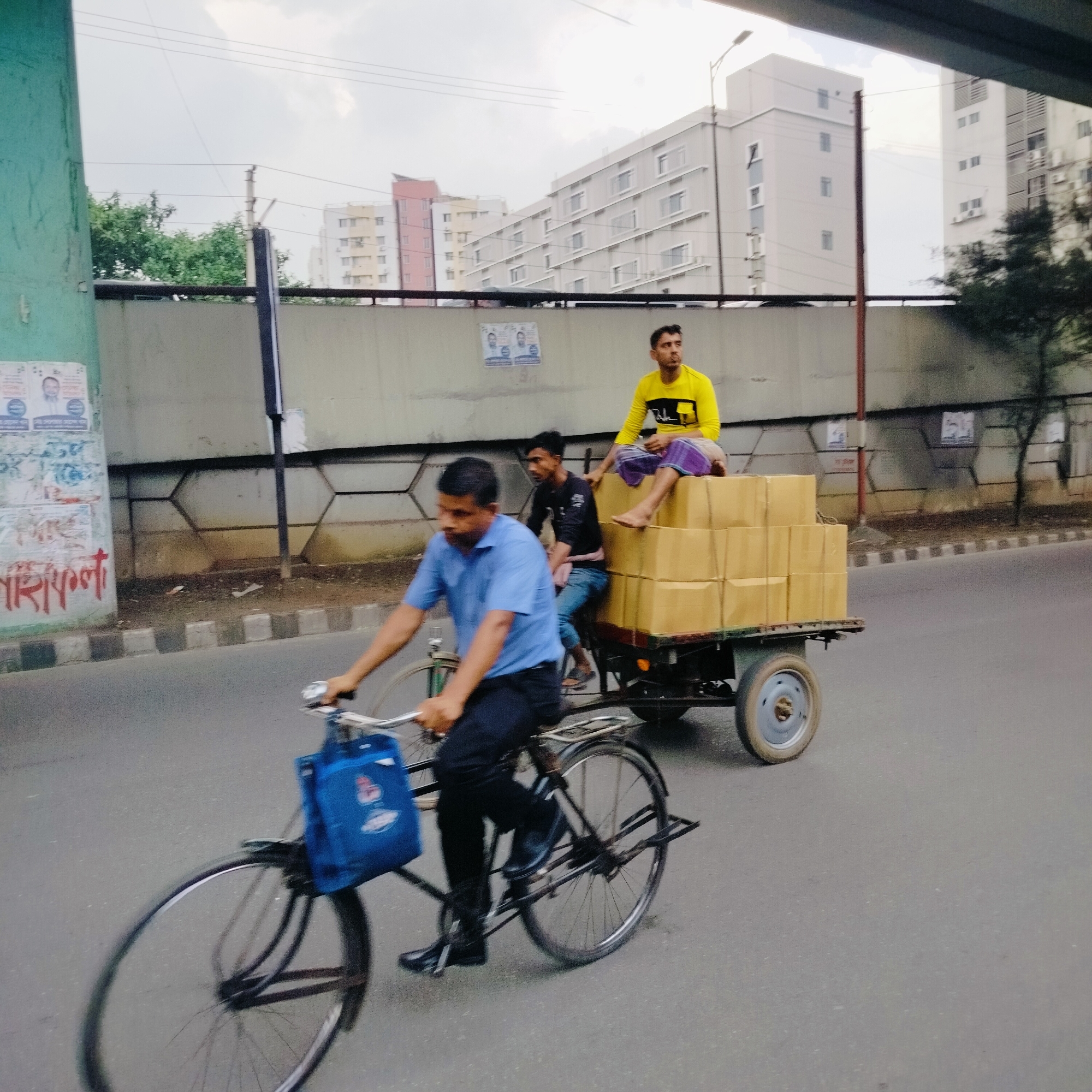 The image depicts a street scene, likely in Dhaka, Bangladesh. A man in a light blue short-sleeved shirt and black pants is riding a bicycle that is pulling a small, two-wheeled cart. The cart is loaded with several tan-colored cardboard boxes. Two other men are on the cart. One is wearing a yellow shirt and a lungi, and is sitting on top of the boxes. The other is wearing a black shirt and blue jeans. The bicycle has a blue bag hanging from the handlebars. The road is paved and there is a curb with a row of grey and white blocks. A concrete wall runs behind the curb. Buildings can be seen in the background. There is an overhead structure partially visible at the top of the frame. The sky is overcast. Graffiti is visible on a green wall.