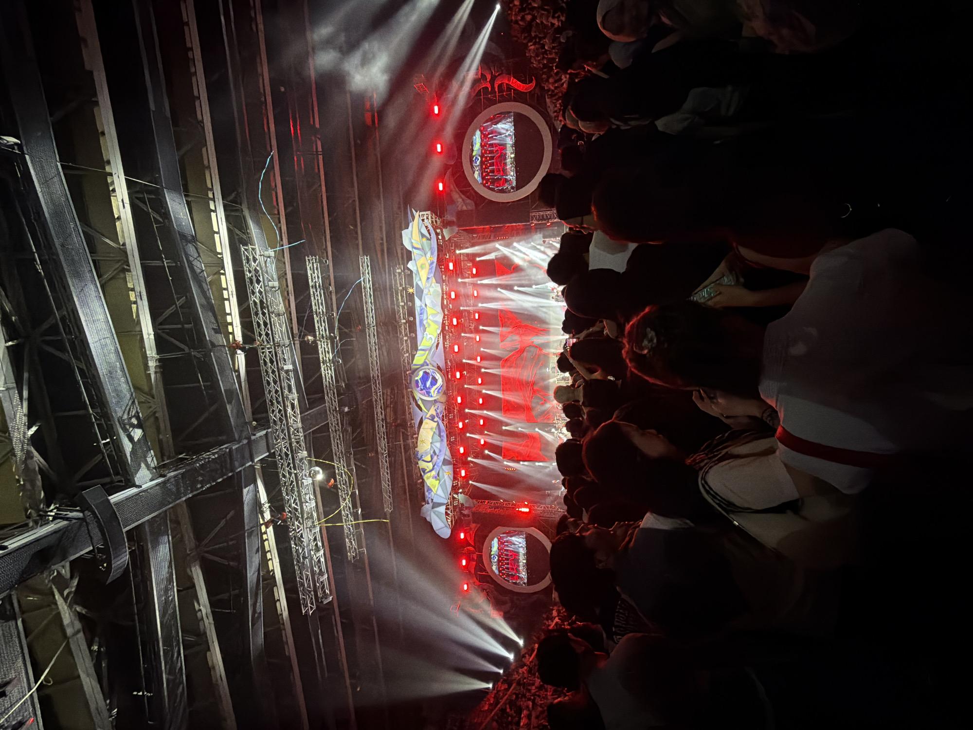 The image captures a vibrant concert scene, with a large crowd gathered in an indoor arena. The stage is elaborately set with bright red and white lights, accompanied by dramatic smoke effects, illuminating the performers and creating a dynamic atmosphere. Two circular screens flank the stage, displaying live feeds of the performance. The ceiling of the arena reveals a complex network of lighting rigs, trusses, and speakers, indicative of a professional setup. The audience, predominantly silhouetted against the stage lights, appears engaged and immersed in the event, with some individuals holding up their phones to capture the spectacle. The overall impression is one of a lively and energetic music performance in a large-scale venue.
