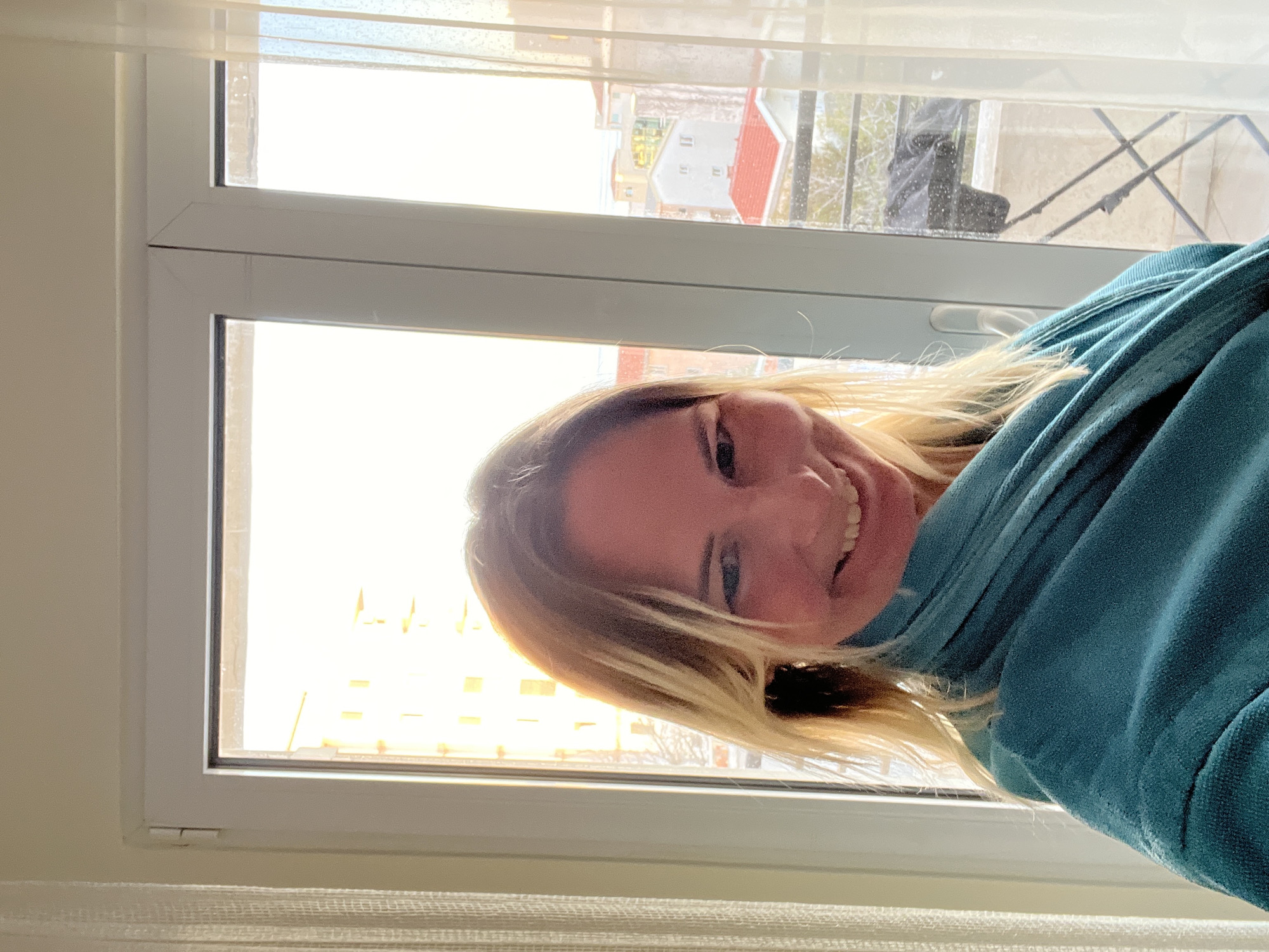 A woman with blonde hair, wearing a blue robe, is looking at the camera from a room with a window. Through the window, the city of Boreti, Montenegro is visible. The window is white framed and slightly open. The room is brightly lit by the daylight. A white curtain is visible at the bottom of the image.