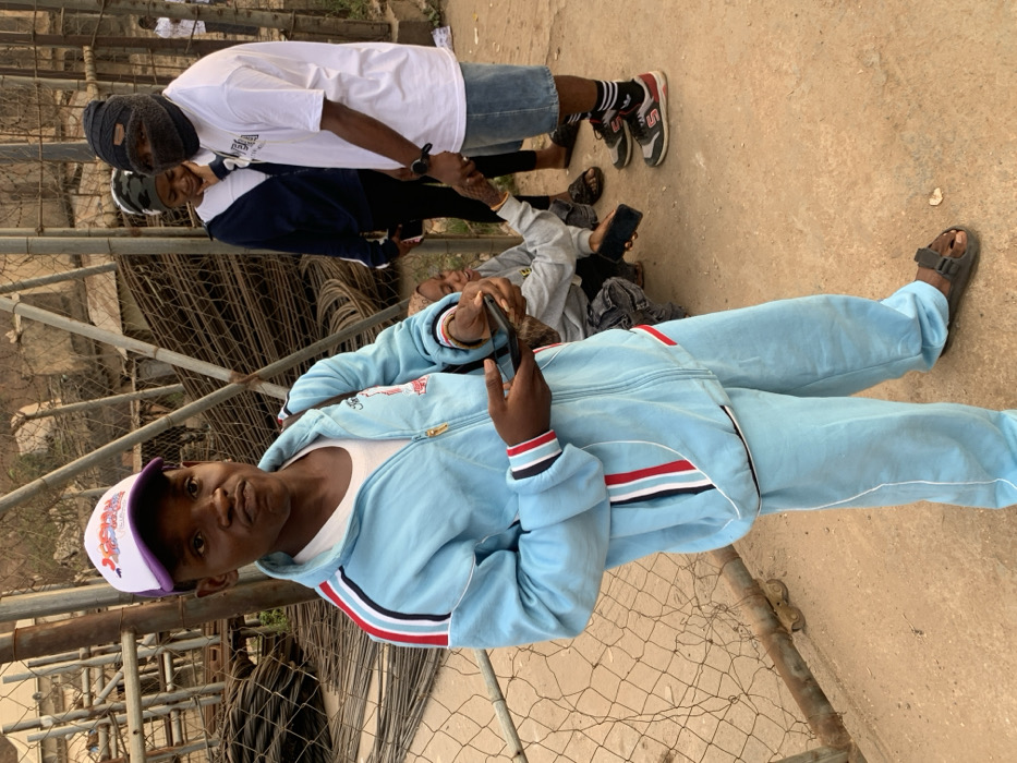 A group of people stands near a chain-link fence. One individual is holding a smartphone, wearing a blue tracksuit and a baseball cap. Another person wears a white t-shirt, denim shorts, and sneakers. A third individual dons a navy and white top. Several other people are visible in the background, some also holding phones. The ground is a mix of dirt and concrete.