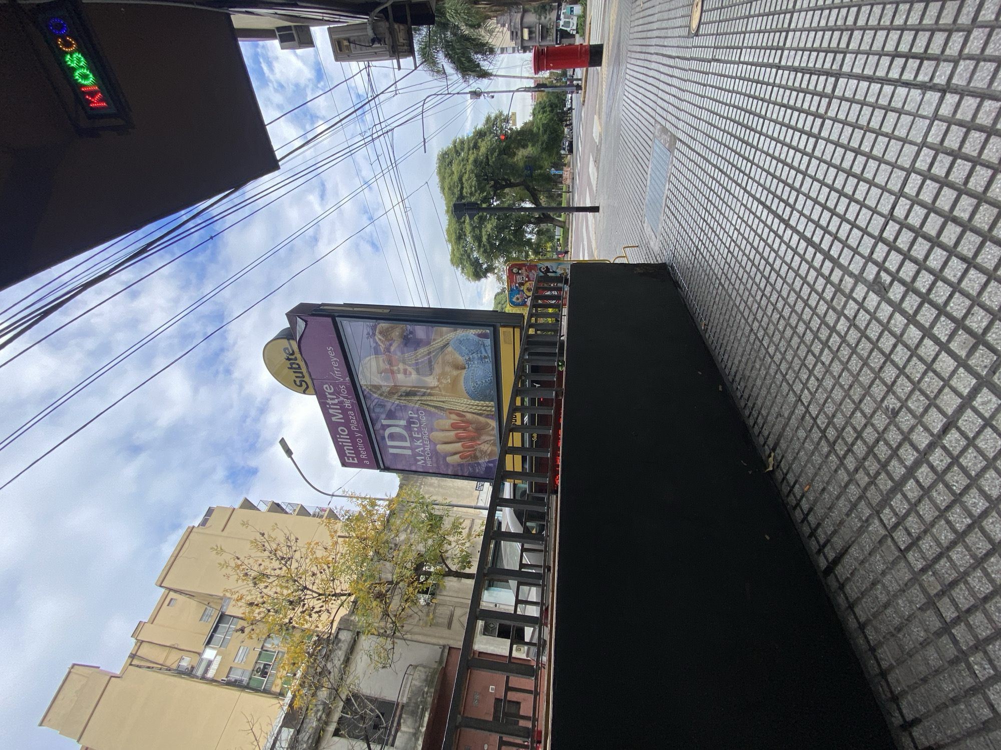 The image shows a street scene in the city of Buenos Aires, Argentina. Visible is a subway entrance with a sign reading "Subte Emilio Mitre a Retiro y Plaza de los Virreyes" with advertising for "IDI MAKE UP HIPOALERGENICO". In the background is a yellow building and bare trees. Overhead are electrical wires and a blue sky with white clouds. The sidewalk has a geometric pattern.