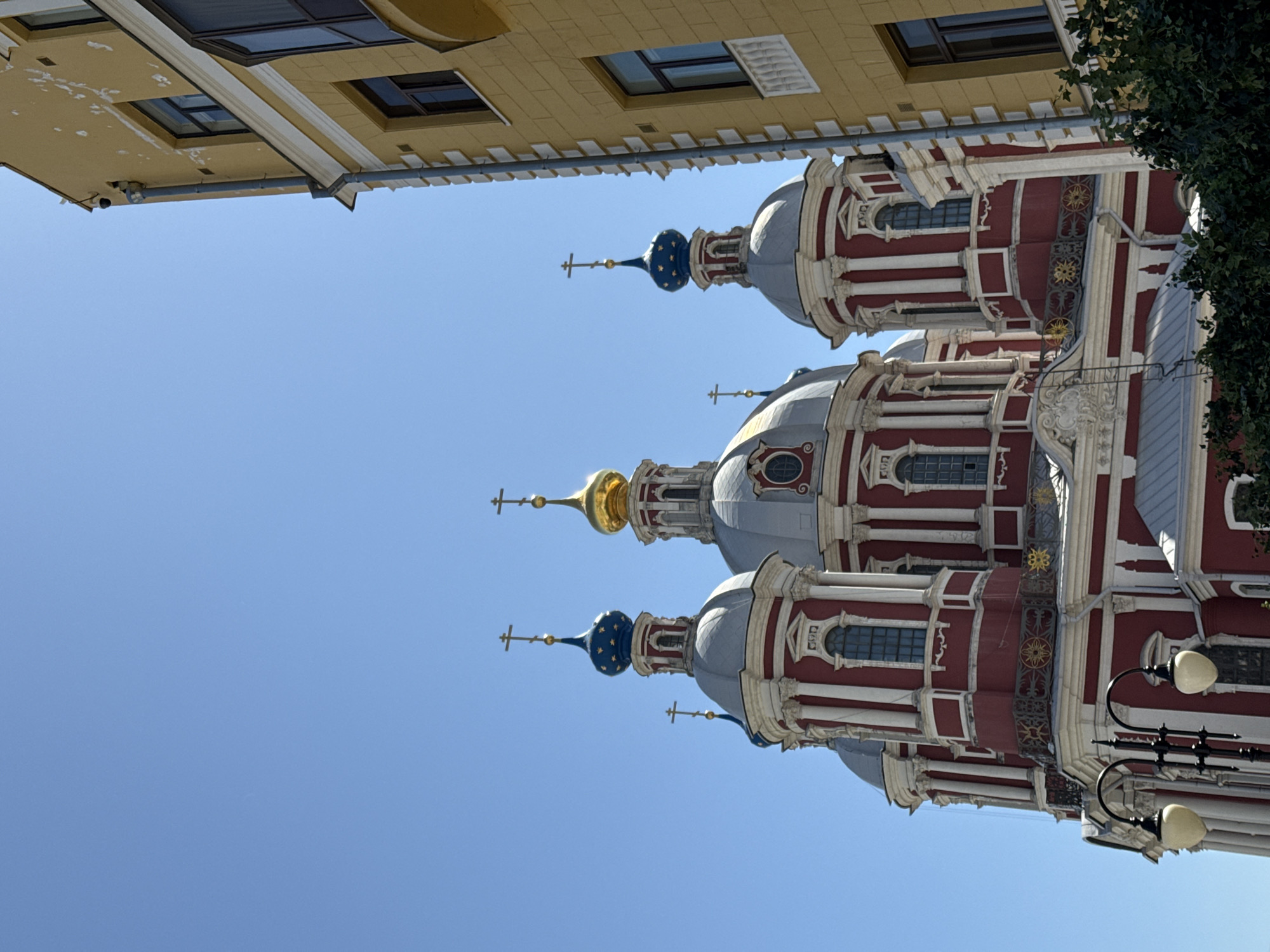 The image shows the Church of the Savior Transfiguration in Moscow, Russia, as seen from a low angle. The church features three domes, two with blue and gold star patterns and one with a golden dome. The building is primarily red and white, with ornate architectural details. Above the church, the corner of a yellow building is visible. The sky is clear and blue. A lamppost is visible on the right side of the church.