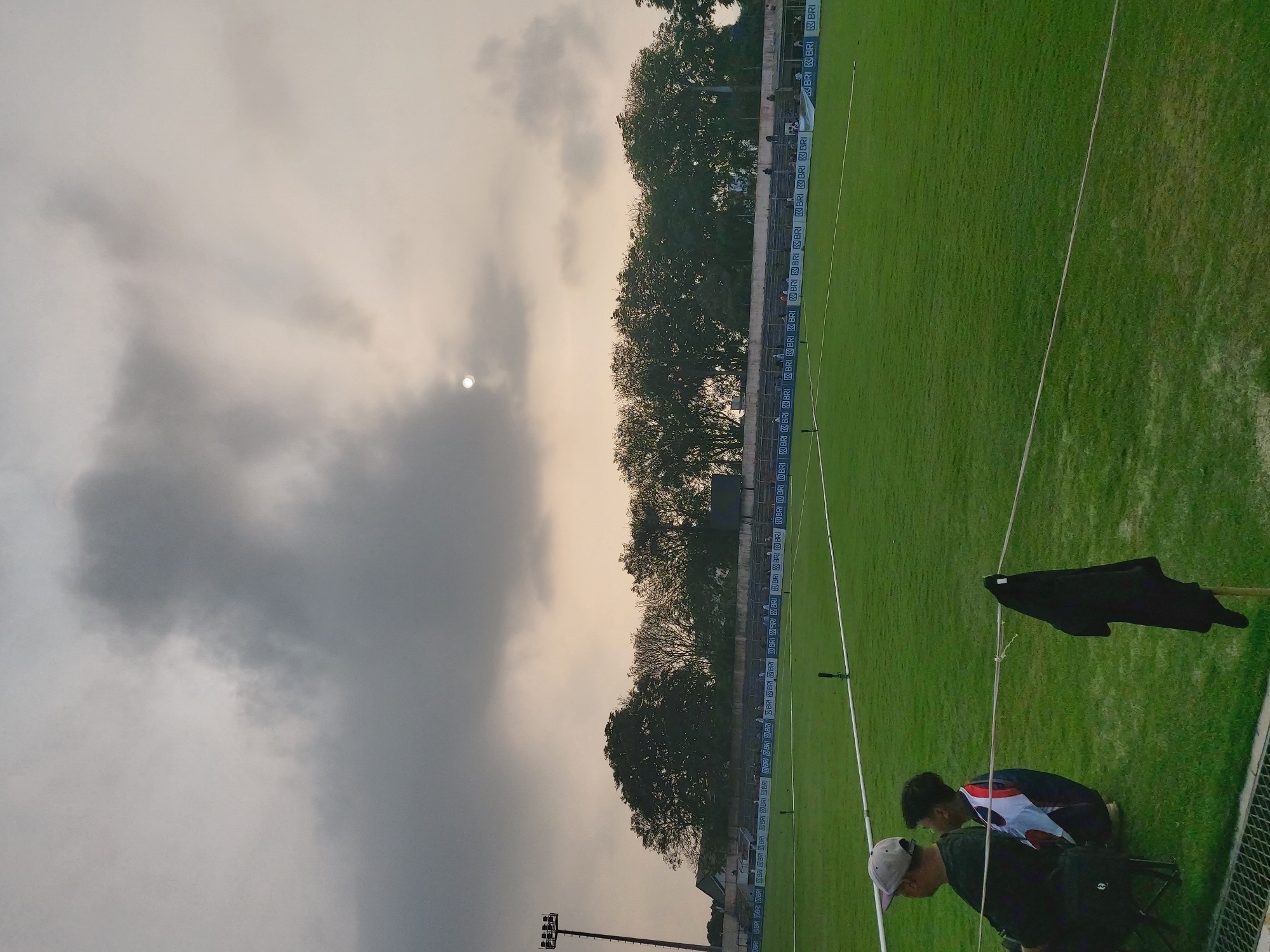 A low-angle, wide shot captures a soccer stadium in Bandar Lampung, Indonesia, under a cloudy sky. The sun is partially obscured by clouds, creating a diffused light. The stadium features a meticulously maintained green grass field with white boundary lines. In the foreground, two people are seated on chairs on the grass, observing the field. One person, wearing a gray cap and dark clothing, is seen from the back. Beside them, a younger person in a colorful shirt is crouched. A black piece of clothing is hanging from a pole in the middle ground. Behind the field, a tiered seating area is visible, with spectators seated and standing. A row of trees forms a backdrop behind the stadium. The stadium also has prominent floodlights. A banner with the logo "BRI" is displayed along the edge of the seating area. The overall atmosphere appears calm and perhaps before or after a sporting event.