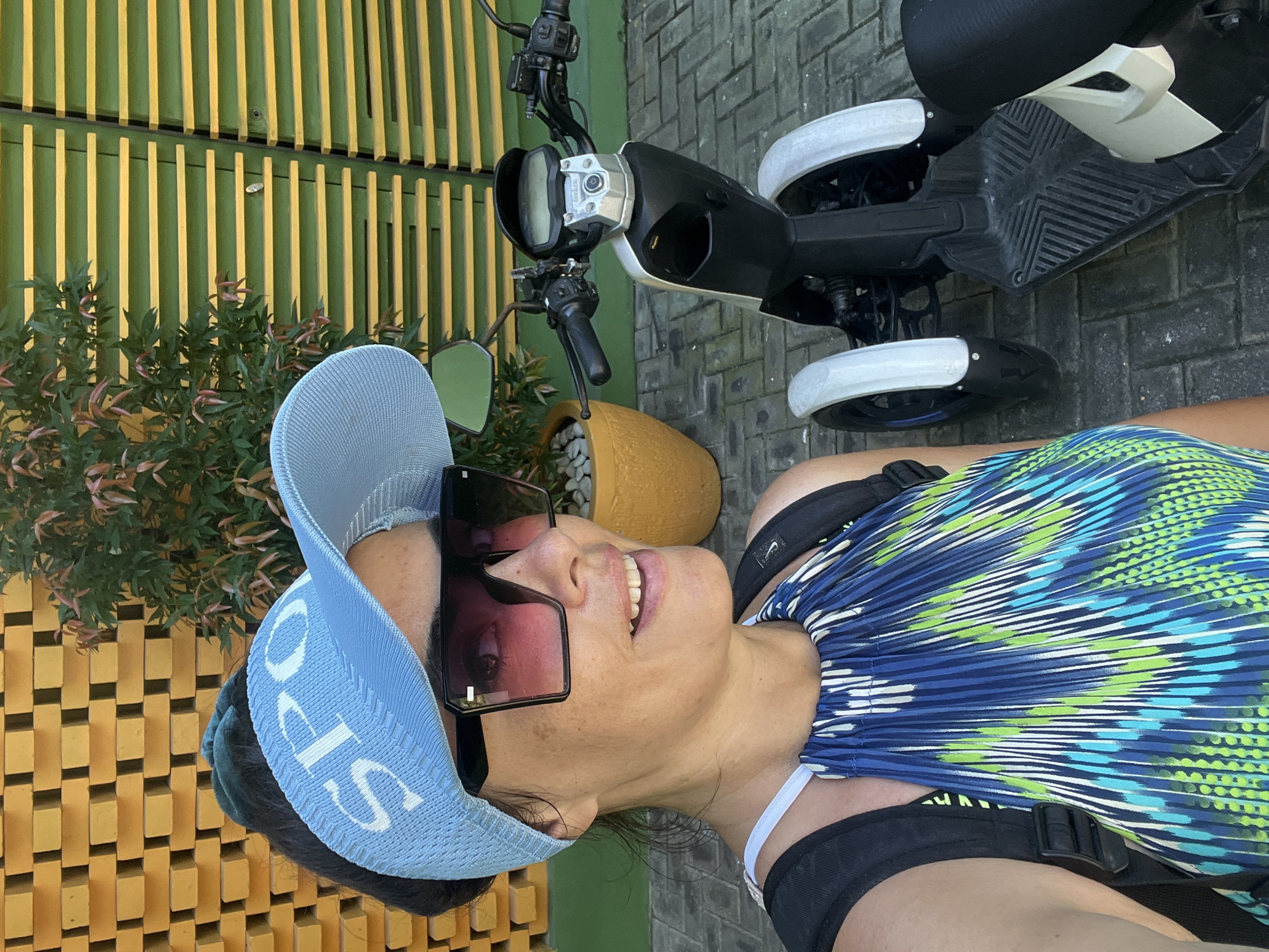 This is a portrait-style photograph of a woman in Kuta Selatan, Indonesia. The woman is wearing a blue and green patterned top, a light blue visor with white lettering, and large, dark-rimmed sunglasses. She is smiling and looking upwards, with her mouth slightly open. Her hair is dark and appears to be tied back.
She is positioned next to a black and white electric scooter, which is partially visible in the background. The scooter has a sleek, modern design with white wheel covers. The woman is wearing a black harness over her patterned top.
The setting appears to be outdoors, possibly on a paved area with brick-like patterns. Behind the scooter and to the left of the woman is a distinctive wall or fence made of alternating yellow and green wooden slats. In front of this slatted wall is a leafy green plant with some reddish-brown new growth. A large, light orange pot containing white stones is also visible near the scooter.
The lighting suggests it is daytime, likely sunny given the bright colors and the shadows. The overall mood conveyed by the woman's expression and the outdoor setting is cheerful and relaxed. There is some visible text on the woman's visor which appears to be white lettering, possibly "SPO".