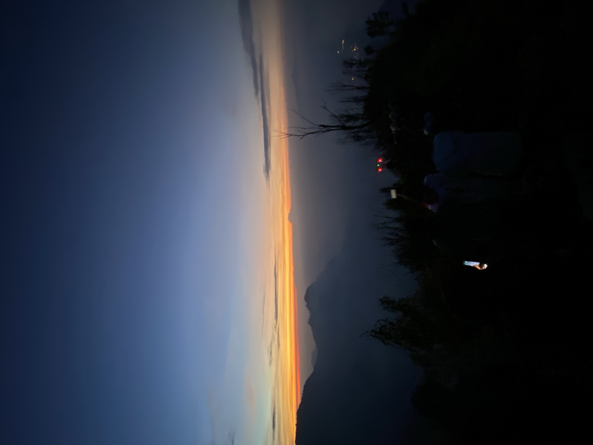 From a high vantage point in Cemorolawang, Indonesia, the scene captures a dusky landscape at the edge of a mountain. The sky transitions from dark blue to a vibrant orange and yellow horizon. The dark silhouette of a mountain range is visible beneath the colorful sky, partially obscured by a haze. People appear on the right side of the frame on top of the mountain, partially illuminated by lights. One person is holding a phone, taking a photo, and another one is holding a flashlight. The foreground is dark, with vague shapes suggesting vegetation. The image is oriented vertically.