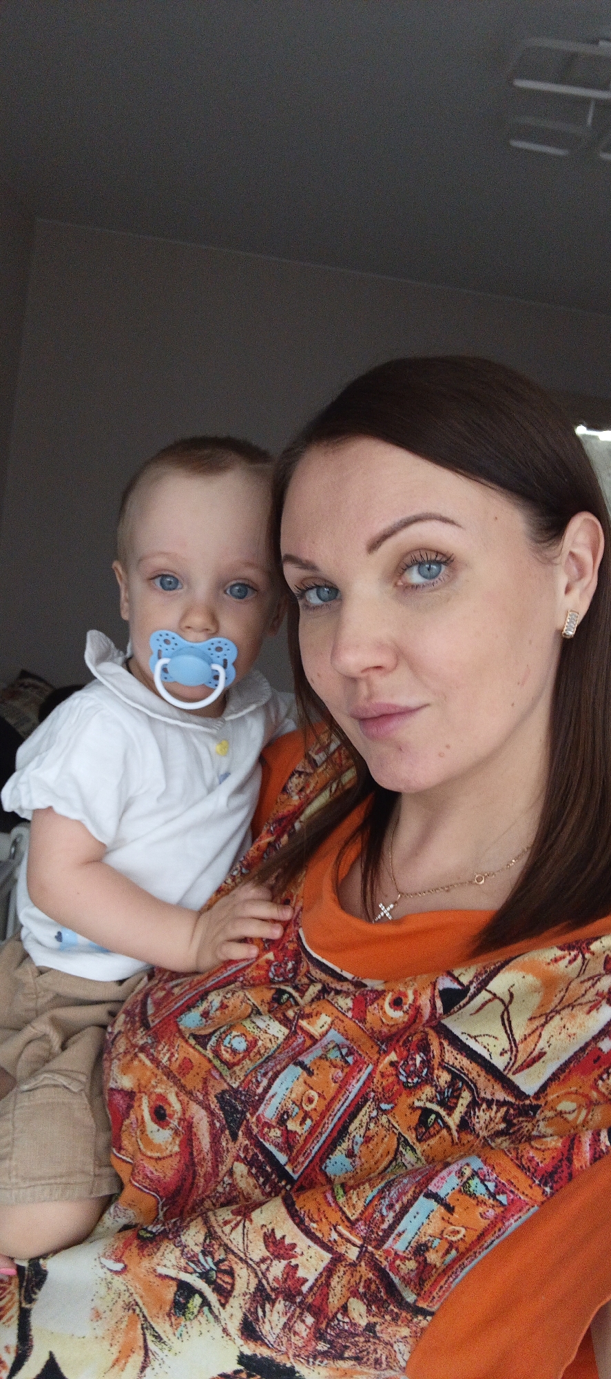 This close-up selfie features a woman holding a baby indoors. The woman, likely the mother, has long, straight brown hair and striking light blue eyes. She wears a vibrant, intricately patterned top dominated by orange, red, and blue geometric and floral designs. A gold-colored necklace with a small cross pendant is visible around her neck, along with another delicate chain, and a small, gold-toned earring with clear stones in her right ear. Her expression is neutral and composed as she looks directly at the camera.
The baby, held in the woman's arms, also has light brown hair and bright blue eyes, matching the woman's. The baby is wearing a white short-sleeved collared shirt and light brown or khaki shorts. A prominent bright blue pacifier, featuring a butterfly or flower-like design, is in the baby's mouth, giving the baby a calm demeanor. One of the baby's hands rests gently on the woman's shoulder.
The background is a plain, light gray wall, suggesting an uncluttered indoor setting, likely a home in Chaikovsky, Russia. The lighting is soft and ambient, characteristic of an indoor environment. No text is visible in the image. The scene captures a personal moment between mother and child.
