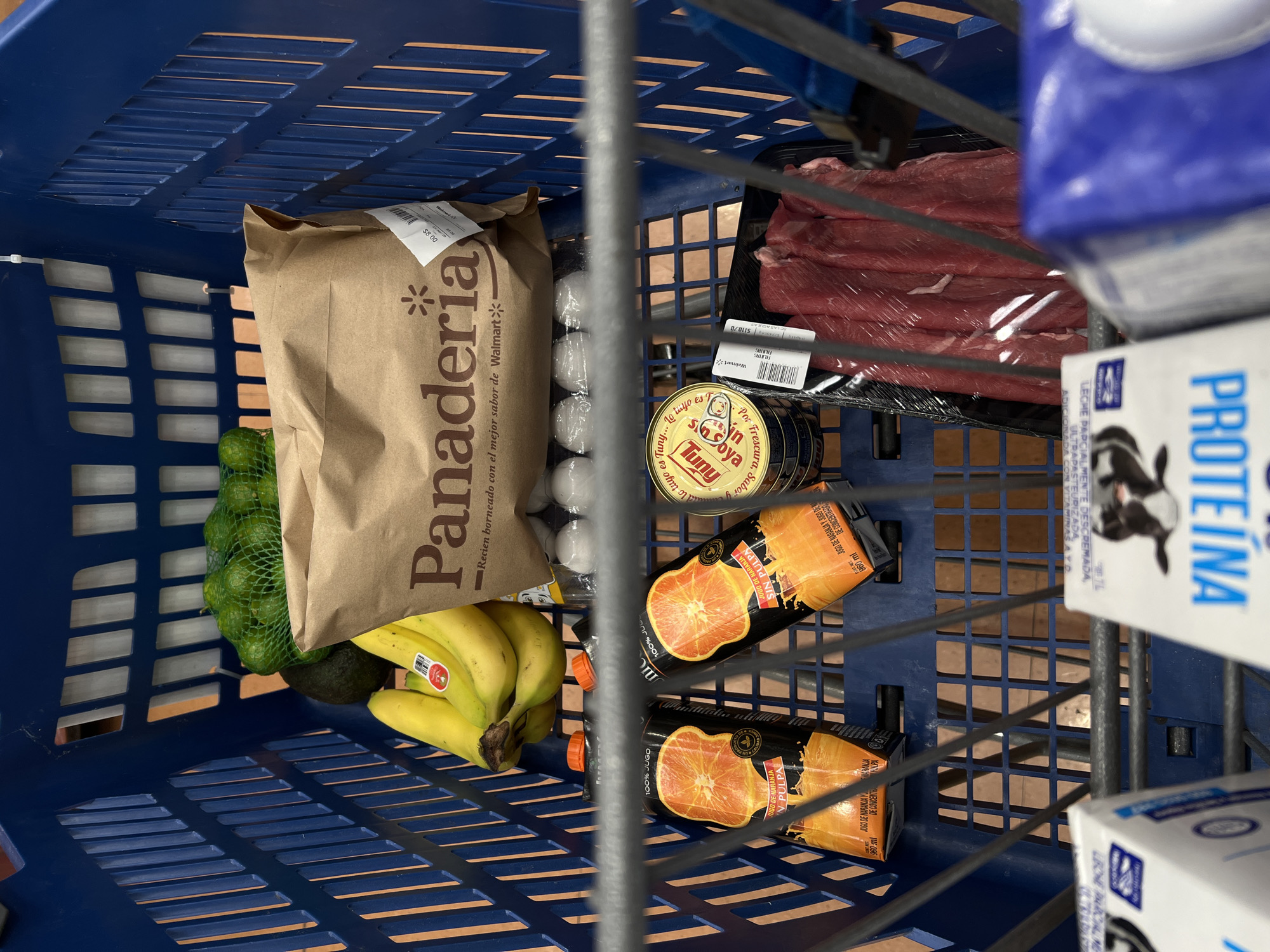 An overhead shot captures the interior of a blue shopping cart, filled with a selection of groceries from a supermarket, likely Walmart, in Mexico City, Mexico. The cart contains a variety of food items.
On the left side, a green mesh bag holds limes, accompanied by a bunch of yellow bananas and a single avocado. A brown paper bag from the 'Panaderia' (bakery) is centrally placed, displaying the text "Recien horneado con el mejor sabor de Walmart" (Freshly baked with the best flavor from Walmart) and a price tag of "$8.00".
Further inside the cart, a carton of eggs is partially visible. To the right, a tray of red meat, possibly beef, is wrapped in plastic film, bearing a 'Walmart' label with a price of "$110/0". Below the meat, several cans of 'Tuny' brand tuna are stacked, with visible text stating "Tuny... Por Frescura, Sabor y sin Soya" (For Freshness, Flavor, and without Soy).
Two cartons of "Jugo de Naranja Sin Pulpa" (Orange Juice Without Pulp) are also present, marked "100% JUGO" and "960 ml". On the far right, partially obscured, are cartons of 'PROTEÍNA' milk, labeled "Leche Parcialmente Descremada, Ultra Pasteurizada, Adicionada con Vitaminas A y D" (Partially Skimmed Milk, Ultra Pasteurized, Enriched with Vitamins A and D). The image depicts a routine grocery shopping activity.