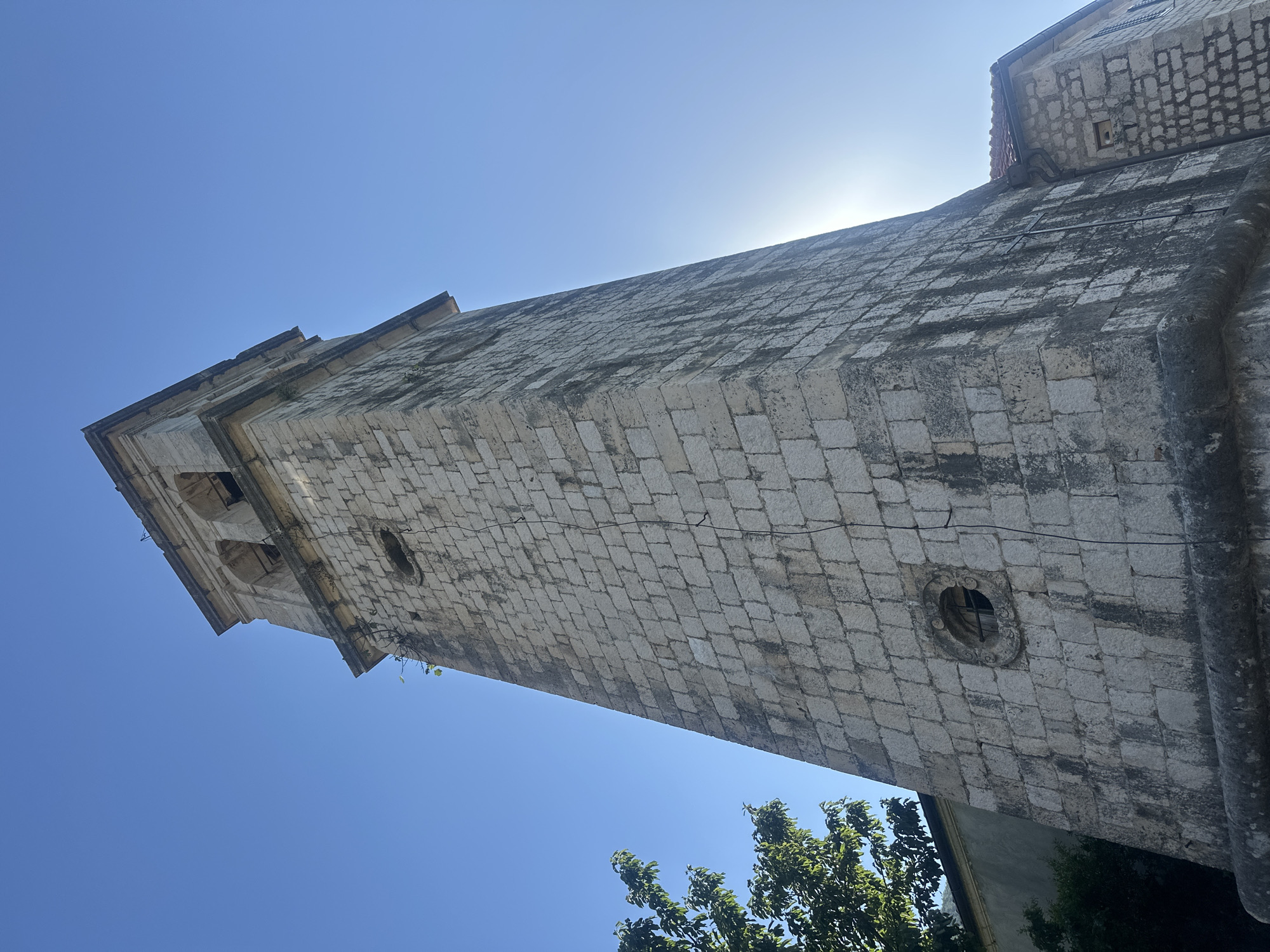 A tall, weathered stone bell tower stands under a clear, bright blue sky. The tower is constructed from light gray, rough-hewn rectangular stone blocks, exhibiting prominent dark vertical streaks indicative of age and moisture runoff. Its base appears slightly flared, and the shaft tapers minimally towards the top. A small, circular oculus with visible metal bars is embedded on the visible side of the tower, approximately midway up. The upper section, a belfry, features two arched openings on the illuminated face, surmounted by a projecting cornice. Strong sunlight emanates from the upper right, producing a distinct lens flare and backlighting the tower, which casts the visible face into a combination of light and shadow, emphasizing its texture. Sparse green vegetation protrudes from cracks in the stonework. To the left, a portion of a leafy green tree is visible. To the right, a section of an adjacent stone building with a terracotta-tiled roof and a dark gutter system is observed. No individuals are present in the scene. This structure is located in Stoliv, Montenegro.