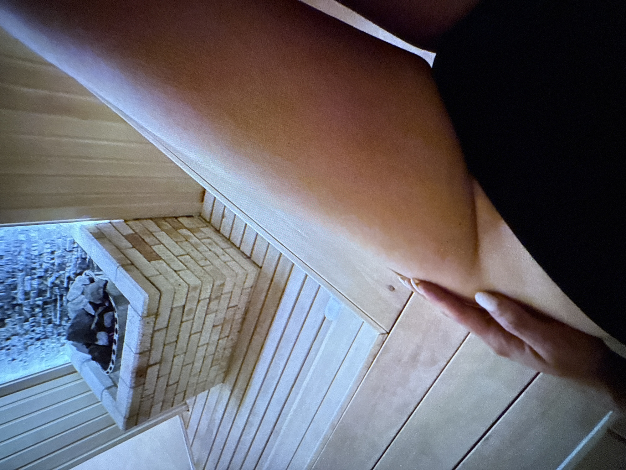 In an indoor sauna, a person's leg and hand are visible. The leg, from the thigh down to the knee, is positioned diagonally across the frame, with the skin appearing smooth and lightly tanned. A hand with neatly manicured white nails rests on the side of the leg, near the knee. The person is wearing dark clothing, visible at the bottom of the frame. The sauna features a wooden bench and floorboards. In the background, a brick structure with stones, likely a sauna heater, is partially visible. The walls of the sauna are lined with vertical wooden planks. The lighting is dim, with a subtle blueish glow emanating from the heater area.