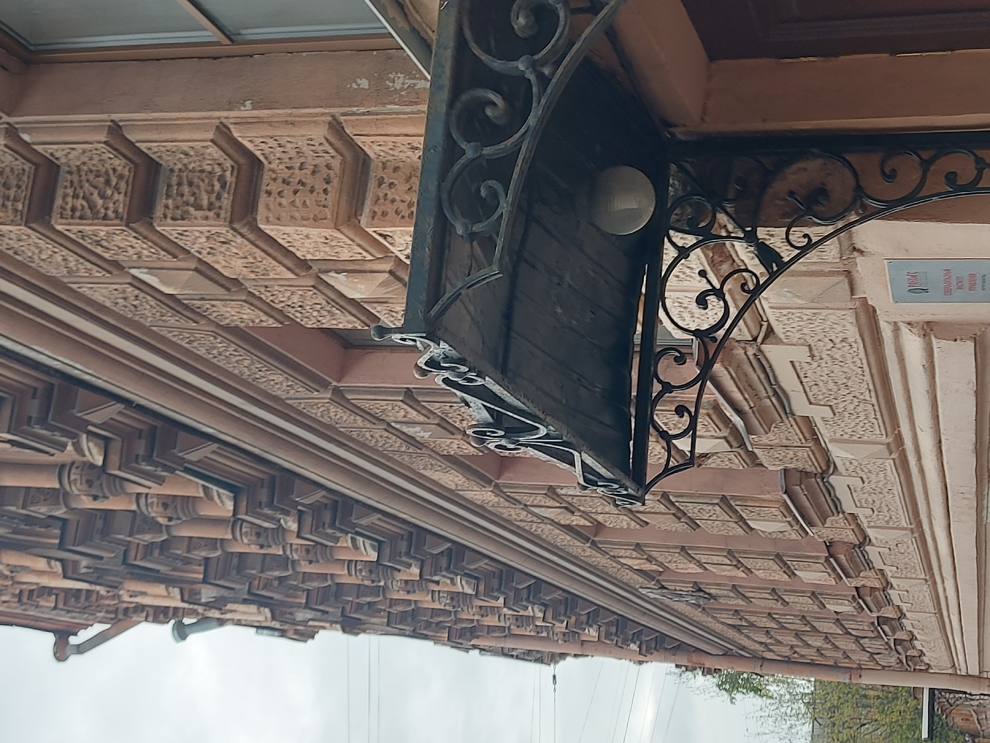 The image depicts the facade of a building in Saint Petersburg, Russia, viewed from a low angle. The building is characterized by elaborate, textured brickwork and decorative molding along the roofline. A wrought-iron canopy, featuring ornate scrollwork and a light fixture, projects from the building's surface. A small sign with text in Cyrillic is visible on the building's wall adjacent to the canopy. A metal pipe runs along the lower edge of the roofline. The sky is overcast, and hints of trees are visible in the distance.
