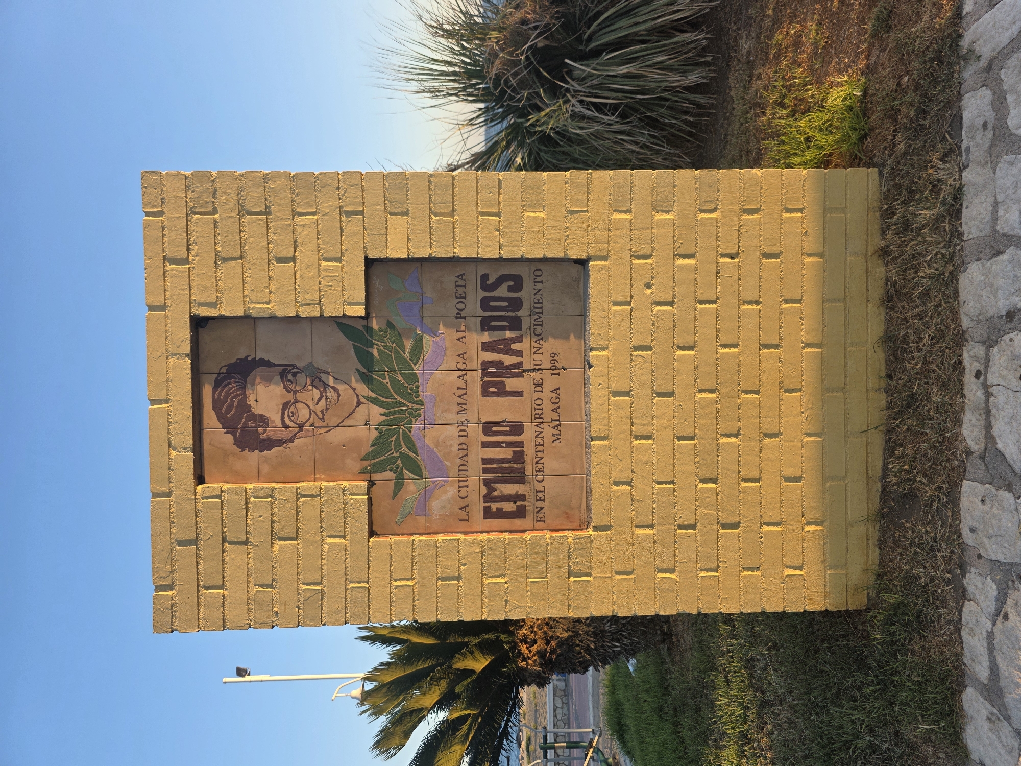 The photograph shows a yellow brick monument with a tiled plaque. The plaque features a portrait of a man with glasses and dark hair, smiling. Below the portrait, text reads: "LA CIUDAD DE MÁLAGA AL POETA EMILIO PRADOS EN EL CENTENARIO DE SU NACIMIENTO MÁLAGA 1999".
The monument is situated outdoors, with a clear blue sky in the background. To the left of the monument, a palm tree is visible, and to the right, a clump of tall, spiky grass. The ground in front of the monument is covered with grass and a paved walkway made of irregular stone pieces. In the distance, a body of water, likely the sea, can be seen. The lighting suggests it is late afternoon or early morning, with long shadows cast by the monument. The overall setting appears to be a public space in Málaga, Spain, possibly a park or a promenade, commemorating the poet Emilio Prados.