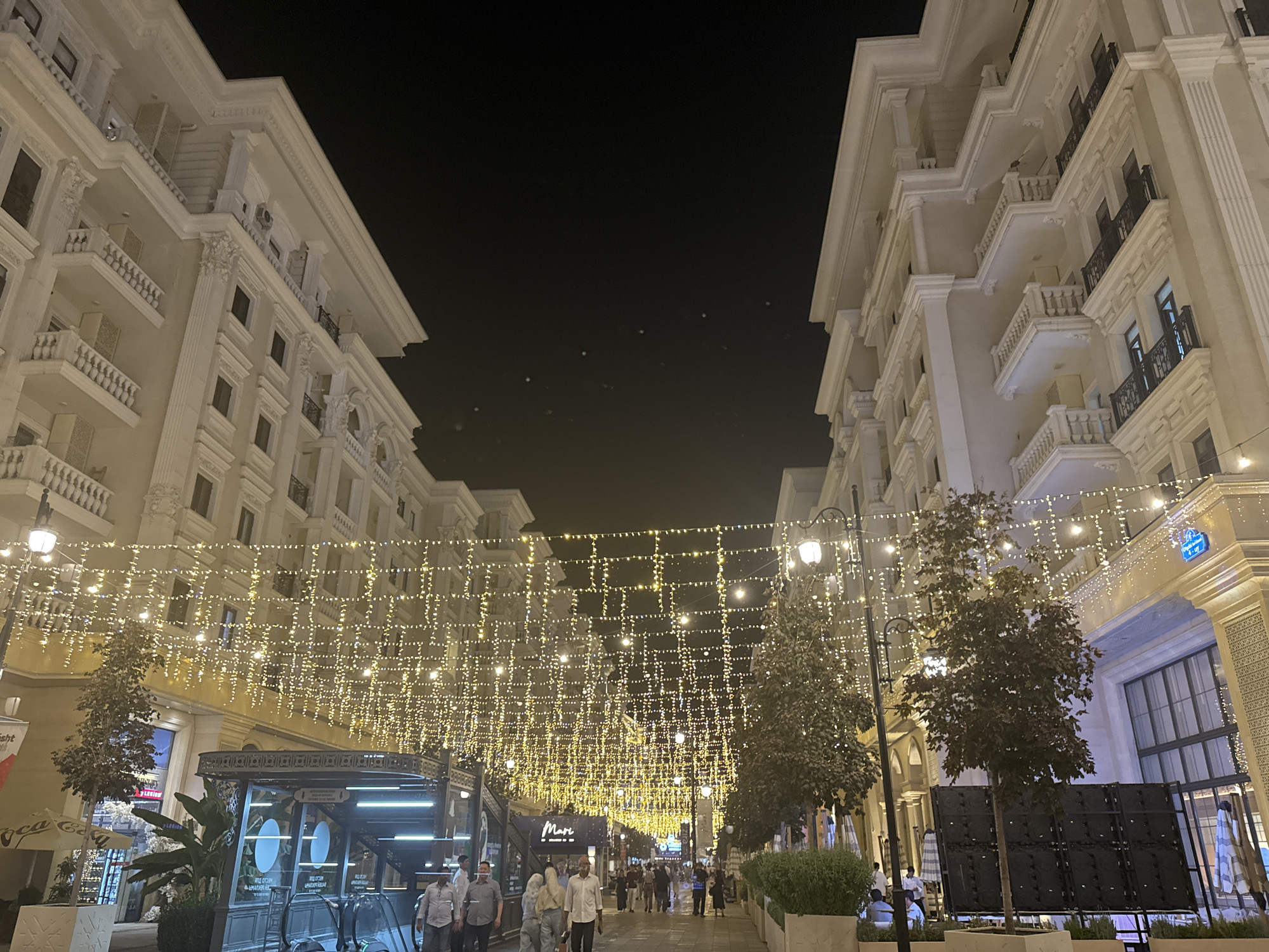 This image captures a vibrant street scene at night in Tashkent, Uzbekistan, likely within a modern, upscale commercial or residential district. The architecture suggests a European influence, with ornate facades of multi-story buildings lining both sides of a pedestrian walkway. The buildings are illuminated, emphasizing their decorative details like balconies with wrought-iron railings and classical columns.
A prominent feature is the extensive string lighting that drapes across the street, creating a festive and somewhat magical ambiance. These lights are densely arranged, forming a canopy of warm, yellow illumination that reflects on the wet pavement, suggesting recent rain.
Several trees, some with broad leaves and others more sparse, are planted in planters along the street, adding a touch of nature to the urban landscape. In the foreground, a contemporary structure with glass elements and signage, possibly an entrance to an underground facility or a modern shop, is visible.
There are people scattered throughout the scene, walking along the street, some appearing to be patrons of the establishments or residents. Their clothing is typical of casual evening wear. The overall atmosphere is one of an active and well-maintained urban environment, showcasing a blend of classical architecture and modern urban design, illuminated for evening enjoyment. The presence of trees and the overall aesthetic suggest an area focused on pedestrian comfort and commercial appeal.