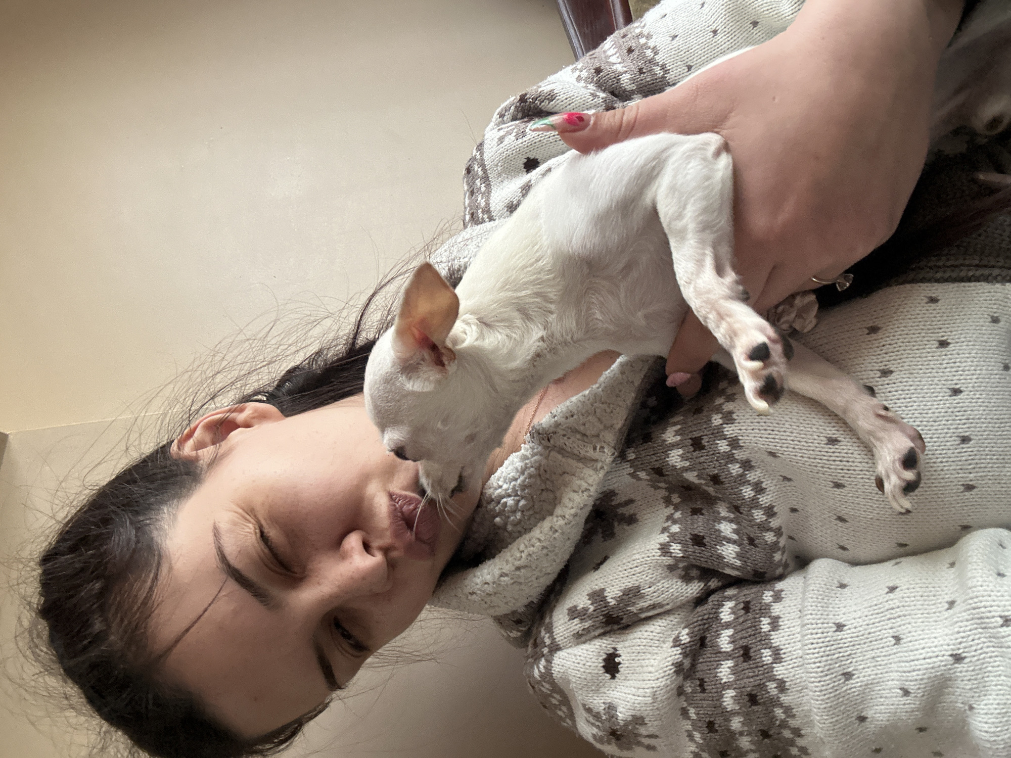 A woman is holding a small, white dog. The dog is licking the woman's face. The woman is wearing a patterned sweater. The location in the city of Sliven, Bulgaria is not discernable from the image.