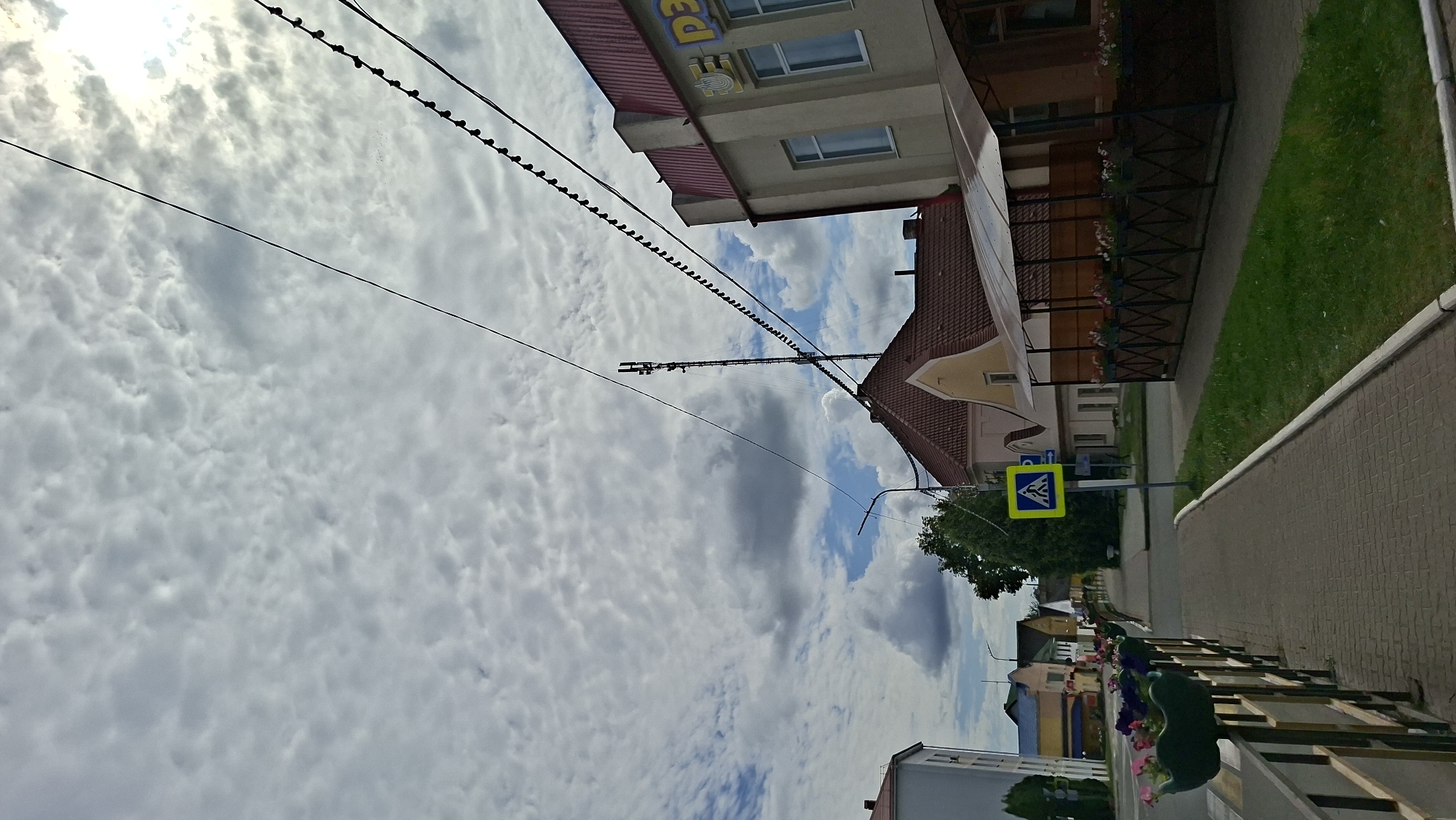 This is an eye-level shot of a sidewalk on a cloudy day in Pastavy, Belarus. The sidewalk is made of brick pavers and is lined with a metal railing and planters filled with pink and purple flowers. Across the street, a yellow pedestrian crossing sign is visible, along with several buildings of varying architectural styles. The building closest to the camera on the right has a red roof and a covered outdoor seating area. In the distance, a telecommunications tower can be seen. The sky is filled with scattered clouds, and power lines crisscross the frame.