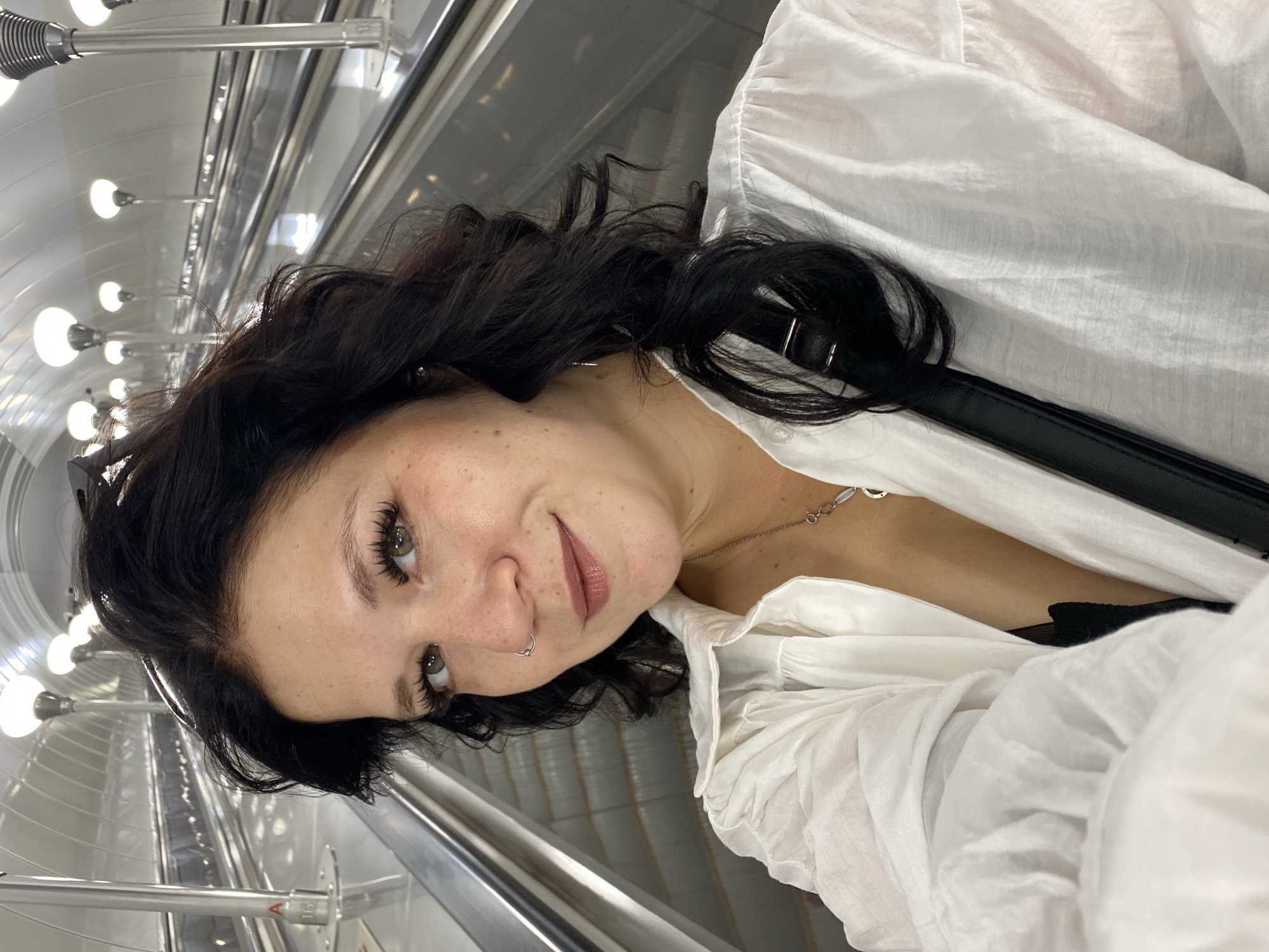 A woman is taking a selfie on an escalator in a metro station in Saint Petersburg, Russia. The woman has dark, wavy hair, brown eyes, and is wearing a white button-up shirt and a black bra. She has a nose ring and a necklace. The escalator is metallic and appears to be indoors. The background shows a curved ceiling with lights and several escalators.