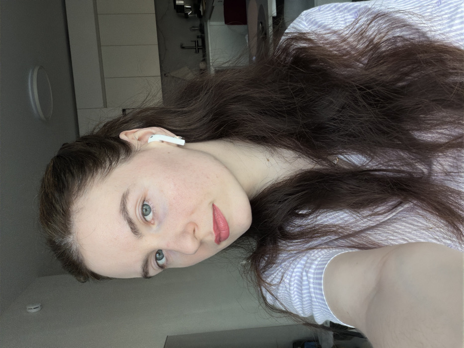 The scene is an indoor portrait of a young adult female within a residence in Ковалёво, Russia. The female subject presents with fair skin, blue eyes, and long, dark brown, wavy hair, partially pulled back from her face. She is wearing pink lipstick and a light-toned, striped collared shirt. A white wireless earbud is inserted into her right ear. The subject directly faces the camera, indicating a selfie. The background consists of a modern kitchen, featuring white upper cabinetry, a stainless steel sink with a faucet, and visible countertop appliances. A circular, flush-mount ceiling light fixture is present. Lighting indicates a combination of natural ambient light from an unseen left source and interior artificial illumination.