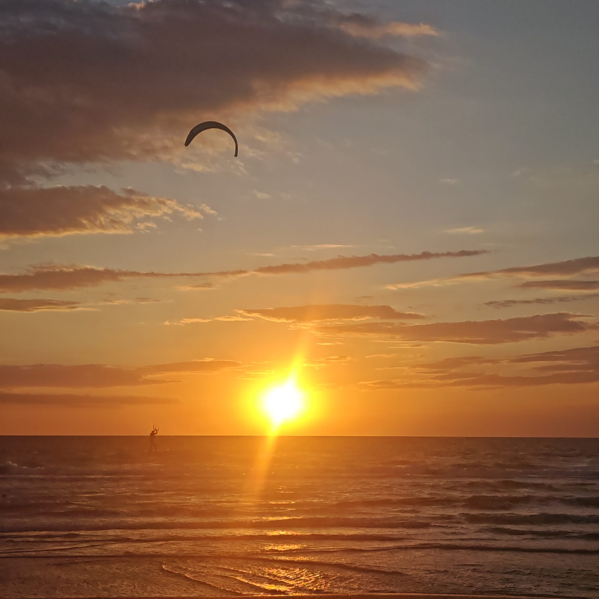 The image shows a sunset over the sea in the city of городское поселение Щелкино, Ukraine. The sun is setting on the horizon, creating a bright glare on the water. In the sky, there are scattered clouds with shades of orange, pink, and blue. A kite surfing wing is visible in the upper left portion of the frame. In the foreground, waves gently roll onto the shore. A distant figure, likely a kitesurfer, stands on the water's surface.