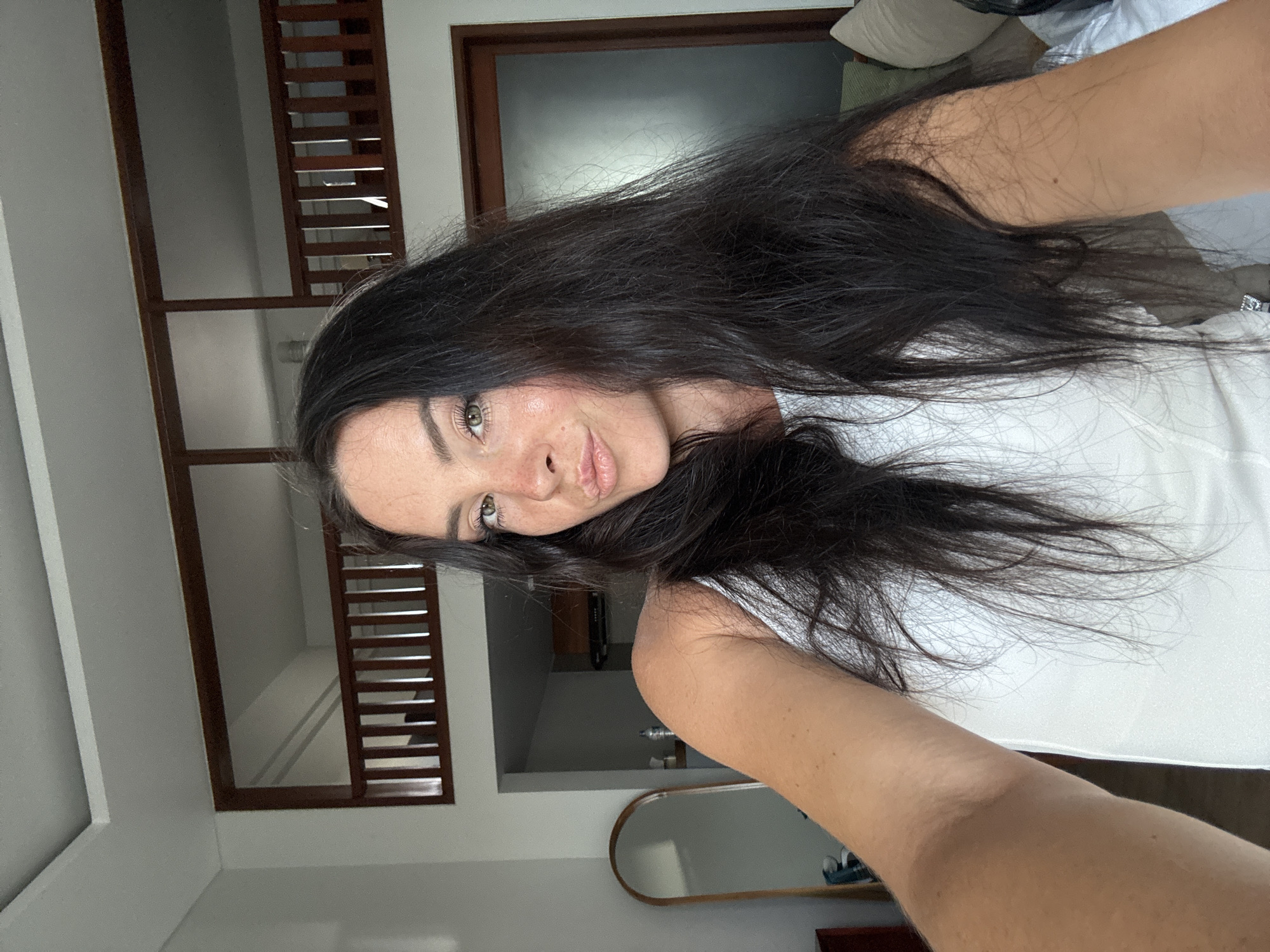 A woman with long, dark hair and fair skin, wearing a white tank top, poses for a selfie in what appears to be a bedroom or living space. She is looking directly at the camera with a slightly pursed-lip expression. Her makeup includes blush on her cheeks and a subtle lip color. The background features a wooden railing structure on an upper level, a large mirror with a wooden frame to her left, and a doorway with a frosted glass panel to her right. The room has white walls and appears to be well-lit, suggesting daytime. Based on the available visual cues such as the architecture and likely tropical setting implied by the lighting and the woman's attire, this scene could be located in North Kuta, Indonesia, a region known for its tourist accommodations with similar interior designs.