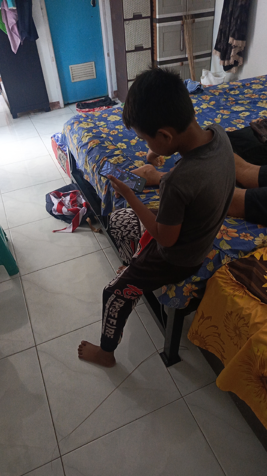 In this indoor scene, a young boy is sitting on the edge of a bed, engrossed in playing a game on his smartphone. The bed is covered with a blue floral-patterned sheet. The boy is wearing a grey t-shirt and black pants with a red and white graphic design. His bare feet are on the white tiled floor.
Behind the bed, a wardrobe with light brown drawers is visible, and to the left, a bright blue door with a vent. A few clothes are hanging on a rack to the right of the wardrobe. A red and white striped fabric is on the floor near the foot of the bed.
The lighting suggests it is daytime, with natural light entering from an unseen source. The overall impression is that of a casual, everyday moment in a home environment, likely in Parung Panjang, Indonesia. The boy's focus on his phone indicates a moment of leisure or entertainment. Text visible on the boy's pants reads "FREE FIRE".