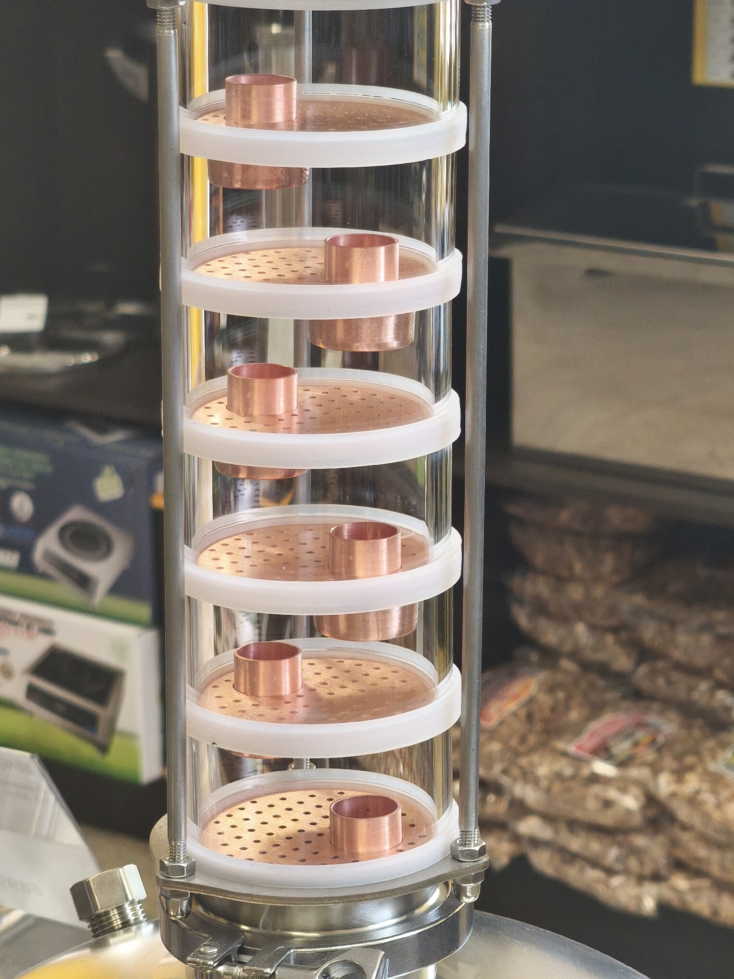 The image shows a close-up of a distillation column with multiple copper trays inside a clear glass cylinder. The column is supported by metal rods and appears to be part of a larger apparatus. In the background, there are out-of-focus elements including what looks like merchandise on shelves, possibly in a store or market setting. The overall impression is of a scientific or industrial piece of equipment. The location in Yoshkar-Ola, Russia cannot be determined from the image.
