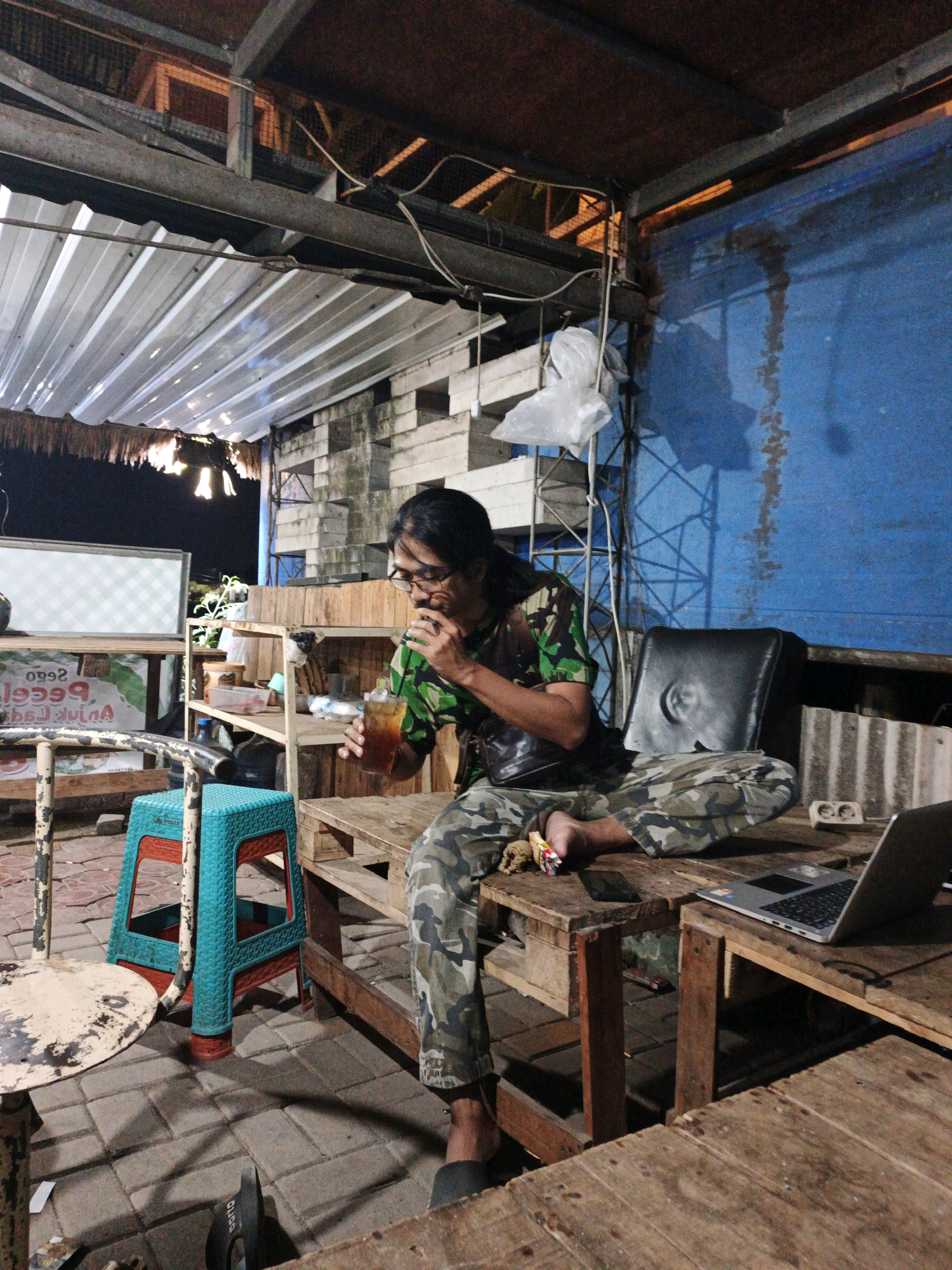 In Ketegan, Indonesia, a person sits at a wooden table outdoors under a corrugated metal roof, drinking iced tea and wearing camouflage clothing. The setting appears to be a casual eatery or rest stop, with a laptop on the table and a blue wall in the background. The ground is paved with interlocking bricks, and a teal plastic stool is visible. A white sign with green checkered patterns is mounted on a shelf behind the person. The lighting suggests it is evening or night.