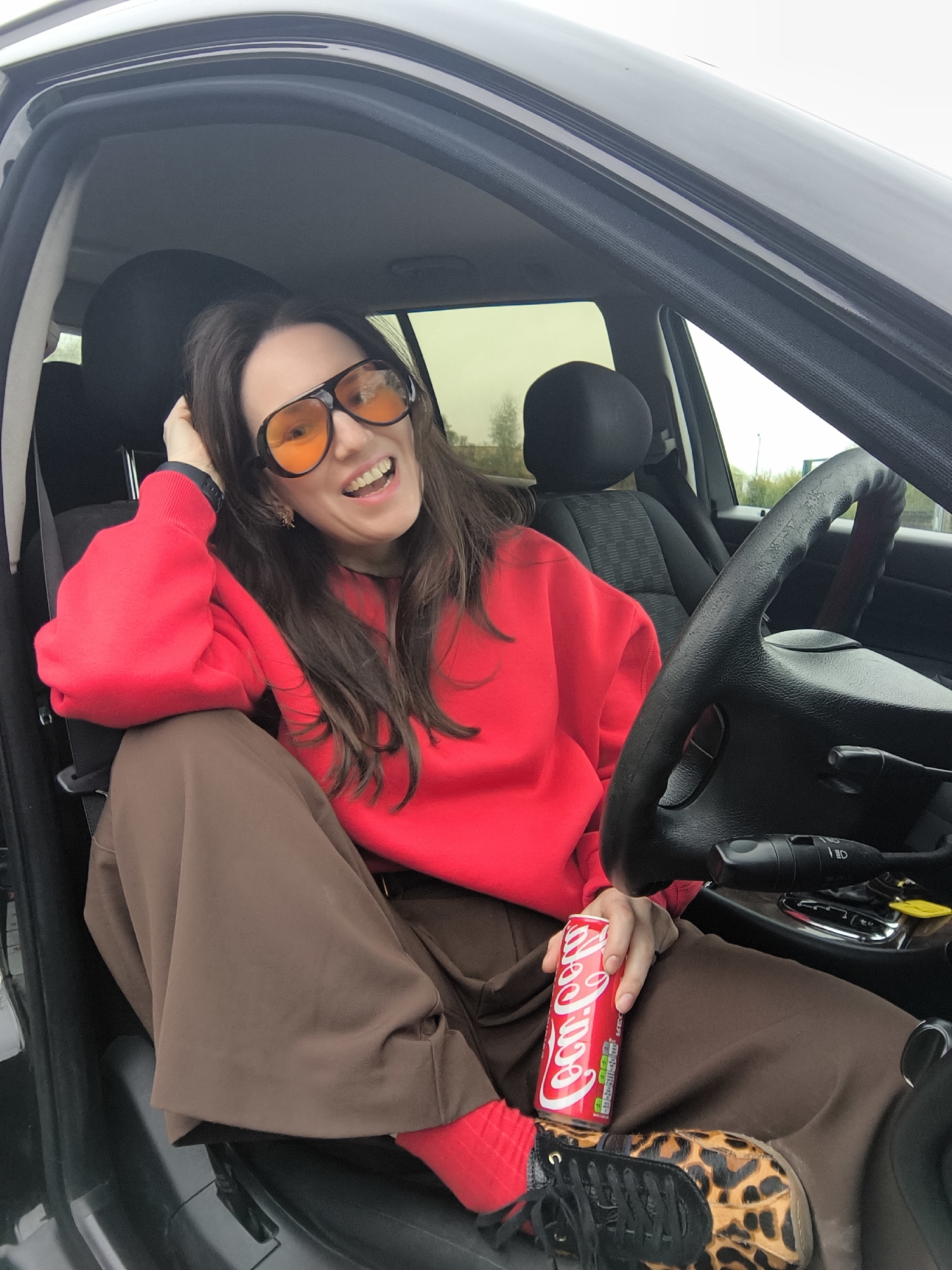 A woman sits in the driver's seat of a car, holding a can of Coca-Cola. She wears orange-tinted glasses, a red sweatshirt, and brown pants. The car's interior is dark, and the steering wheel is visible. One foot shows a shoe with a black laces and a leopard print pattern. The exterior through the car window shows a view of what could be trees or buildings. It is impossible to determine the specific location of Riga, Latvia, from the content of the image.