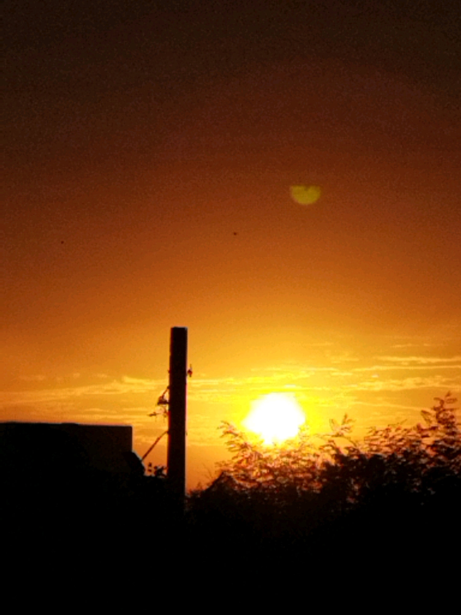 A silhouette of a building and a utility pole are visible against a sunset. The sky is a gradient of deep orange to a lighter yellow. A bright, blown-out sun is low on the horizon, partially obscured by trees and shrubbery. In the upper right portion of the sky, there is a small, faint, heart-shaped halo or atmospheric phenomenon. The image appears to have been taken in Bucharest, Romania, during sunset.