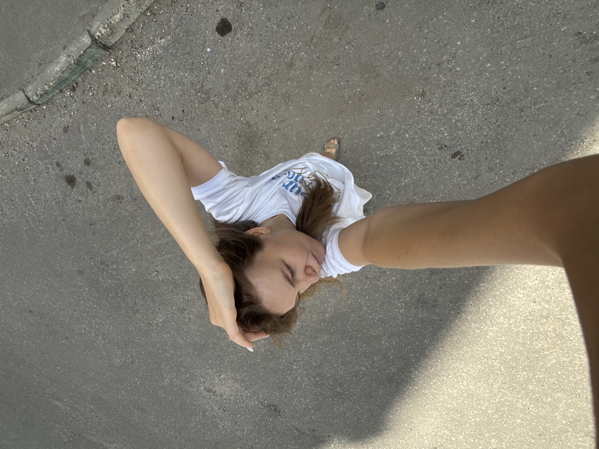 The image shows a young woman from a high-angle perspective, as if she's taking a selfie while standing on a paved surface. She is wearing a white t-shirt with blue text and light-colored sandals. Her eyes are closed, and one hand is resting on her head, suggesting a relaxed or thoughtful pose. The ground beneath her is asphalt or concrete, with some debris and a curb visible on the left side, indicating an outdoor setting, possibly a street or sidewalk in Vladimir, Russia. The lighting suggests it's daytime, with shadows cast by an unseen object on the right. The overall mood is casual and serene. The text on her t-shirt appears to be "YOUNG LOVE" in blue.