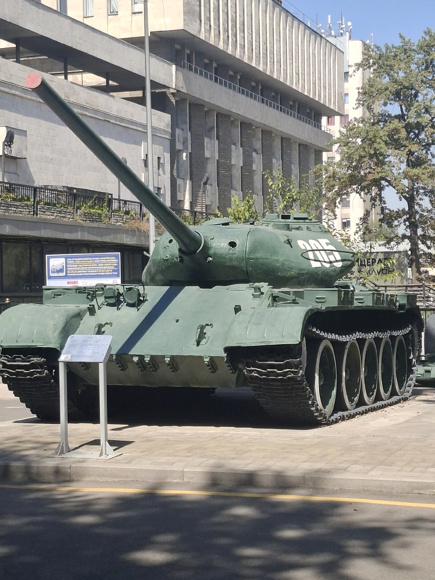 The image shows a green tank on display outdoors, possibly as a war memorial or museum exhibit. The tank is positioned on a paved surface with a yellow line visible in the foreground. In the background, there is a large, modern building with a brutalist architectural style, characterized by concrete facades and protruding elements. Trees with green foliage are also visible, suggesting a temperate climate. The tank itself is heavily armored, with a long barrel and tracks. The number "205" is painted in white on the turret. A sign with text and graphics is mounted on the front of the tank, likely providing information about its history or specifications. The sky is clear and blue, indicating a sunny day. The overall scene depicts a static display in an urban environment. The tank appears to be a Soviet-era model, possibly an IS-3 heavy tank, given its distinctive hull shape and turret. The presence of the building and the urban setting suggest that this is likely a public space within a city. While the image does not explicitly confirm the location as Almaty, Kazakhstan, such displays of military hardware are common in former Soviet republics.