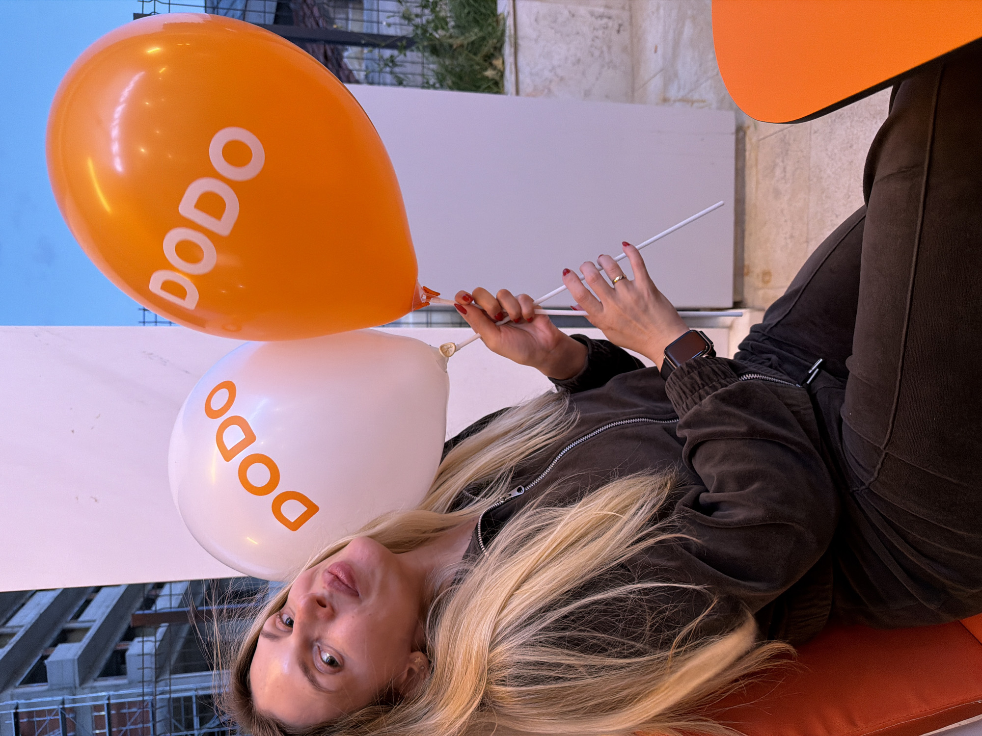 A woman is reclining on an orange cushion, holding two balloons—one orange and one white—each printed with the word "DODO" in white and orange, respectively. She is wearing a dark jacket and dark pants. Her blonde hair is spread out behind her head. She is looking at the camera.
The background includes a white wall, the sky, and the silhouette of a building under construction. The photo was taken in Budva, Montenegro.