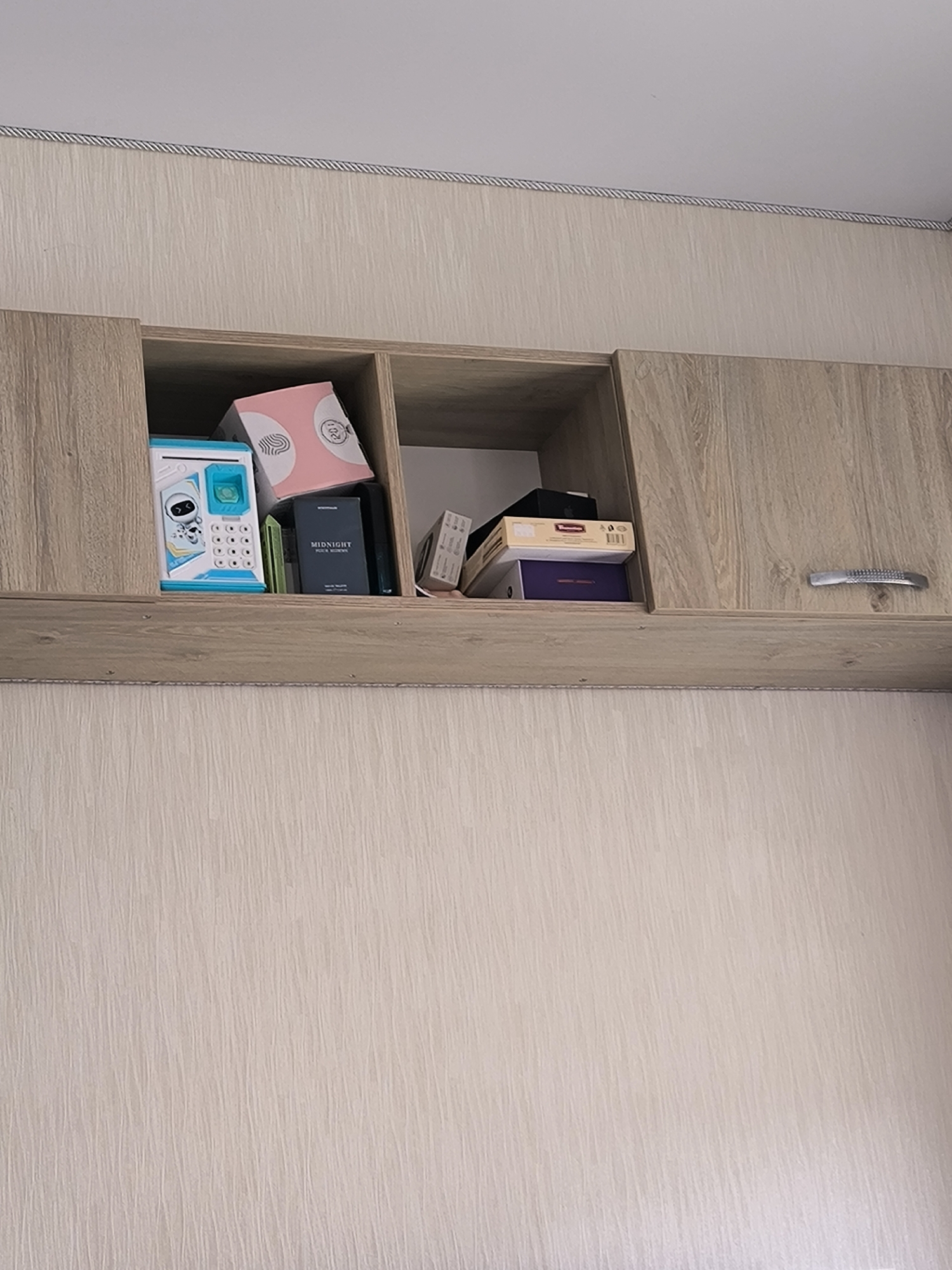 The image shows a top view of a light wood laminate cabinet with a silver textured handle on the right-hand door. Inside the cabinet, there are two compartments. The left compartment contains a blue and white piggy bank shaped like a robot, a pink box with a white swirled pattern, and a dark blue box with "MIDNIGHT" printed in white. The right compartment contains several stacked boxes, including one with a red and yellow label and a dark purple box. The background is a textured light beige wallpaper. There are no people or animals visible in the image, and no actions or interactions are depicted. The scene appears to be an indoor domestic setting, likely a bedroom or living area. The location in Chisinau, Moldova cannot be determined from the visual information provided.