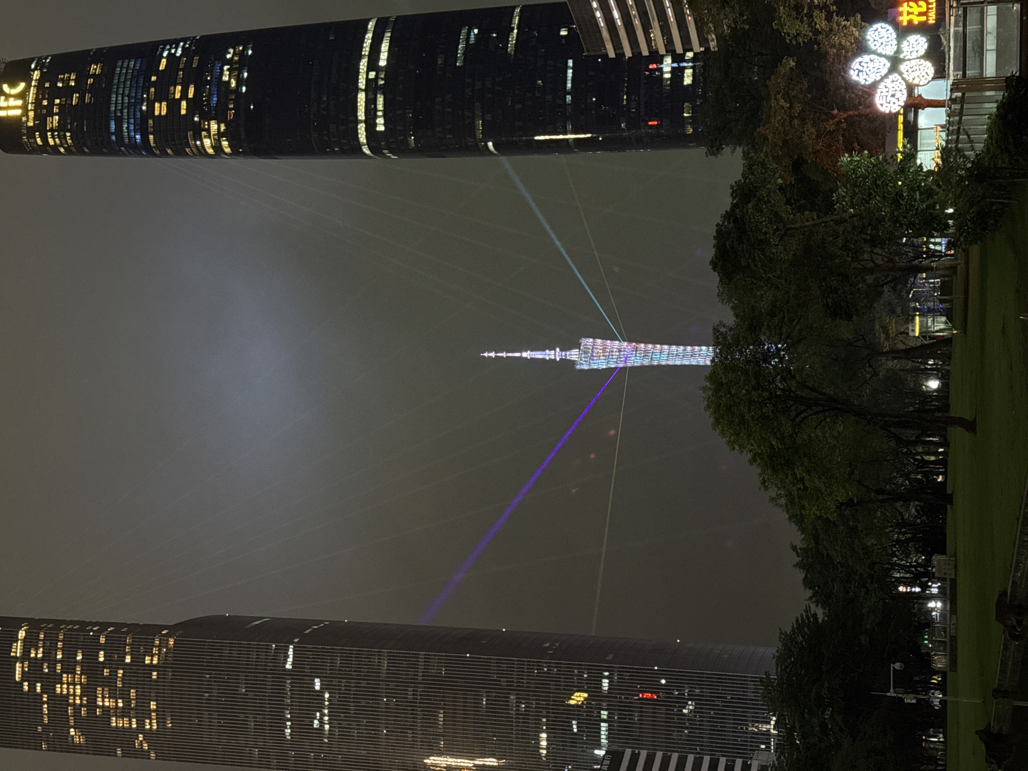 The image depicts a nighttime scene in the Tianhe District of Guangzhou, China. Prominently featured is the Canton Tower, illuminated with colorful lights and intersected by blue and white laser beams. The cityscape includes tall buildings with visible window lights, including the Guangzhou International Finance Center (IFC) with "IFC" signage. Lush trees line the scene's edge, bordering a grassy area. Additional lighting fixtures and possibly signs are seen amidst the trees. The overall environment suggests a humid, possibly hazy night.