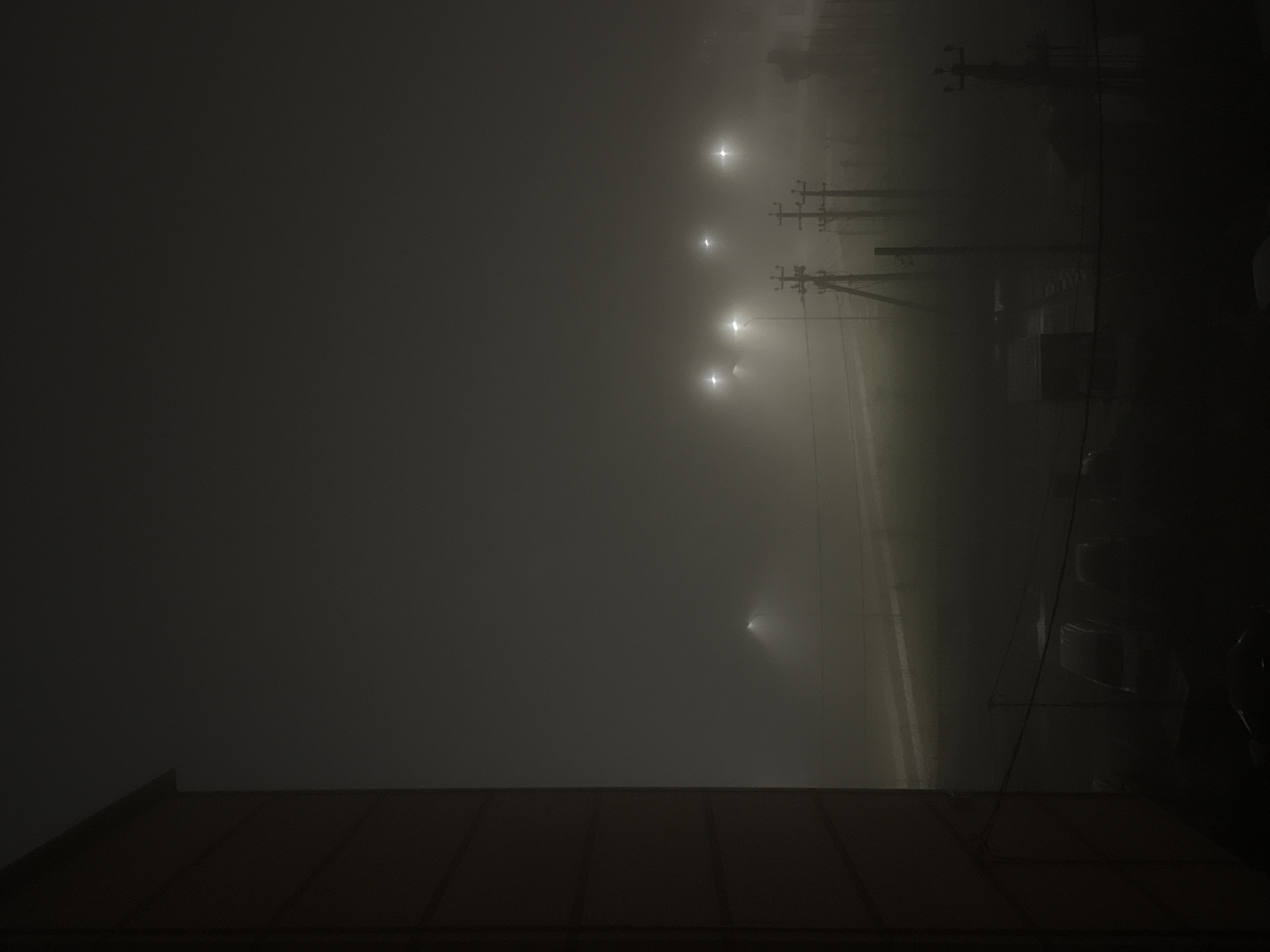 A horizontal, outdoor, low-angle shot depicts a foggy, dimly lit scene in an urban area. In the foreground, the dark roof of a building is visible. Further into the distance, a road runs alongside a row of evenly spaced streetlights. The fog obscures the visibility of most details. Overhead power lines stretch out from the streetlights, appearing as thin black lines. The lights emit a faint glow that diffuses into the heavy fog. No people are visible in the scene.