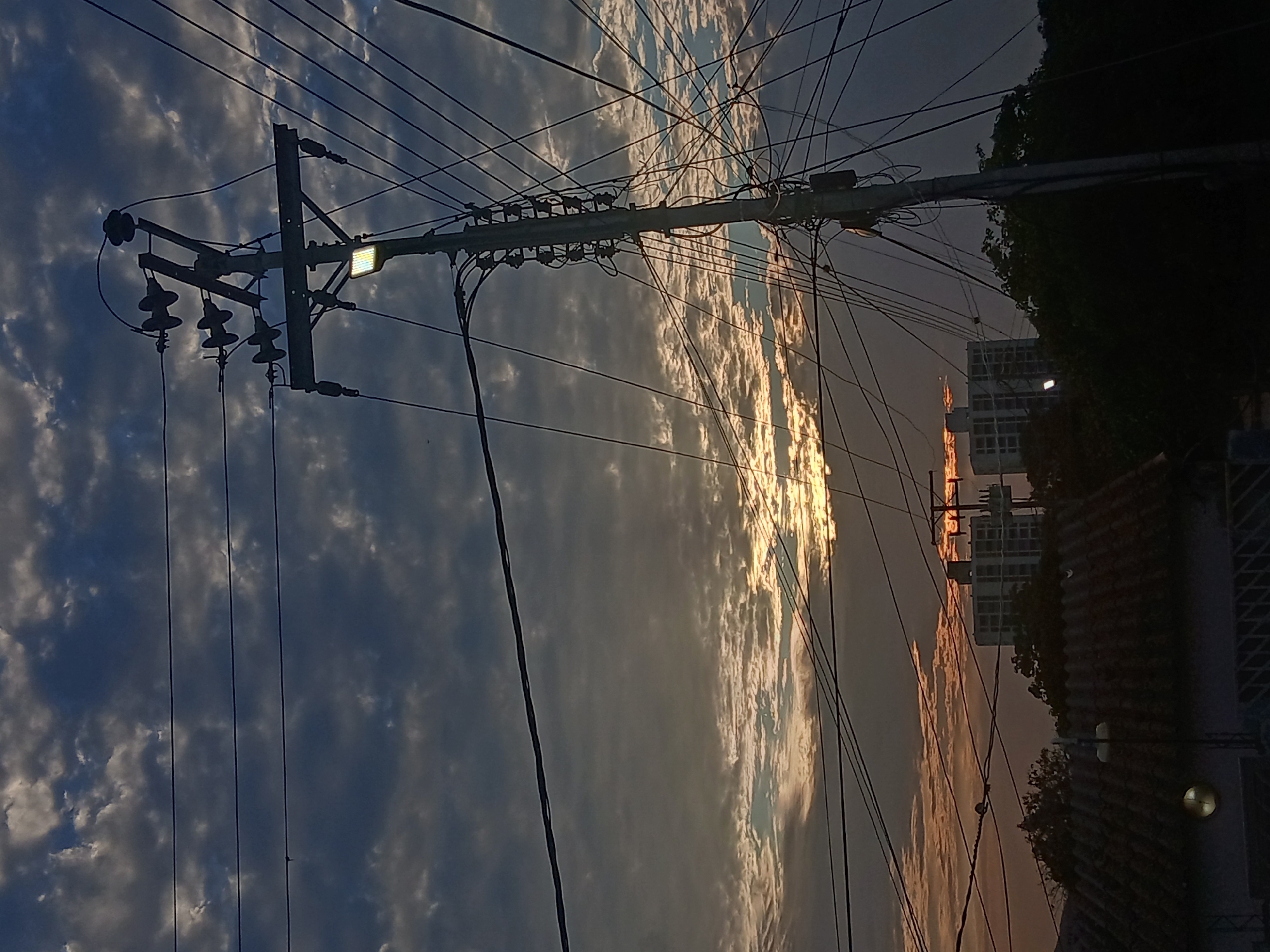 A low-angle, rotated view captures a utility pole in Juan Griego, Venezuela. The pole is on the left side of the image and is connected to many power lines running across the scene. The sky is overcast with clouds, and the sun is attempting to break through. The sky contains gray clouds on the left and warm, sunny clouds on the right, suggesting the time of day is near sunset. The power lines are dense and tangled. Buildings are visible in the background, obscured by the power lines and trees. The scene is likely captured near a residential area with visible street lighting. The overall atmosphere is a mix of dusk and urban infrastructure.