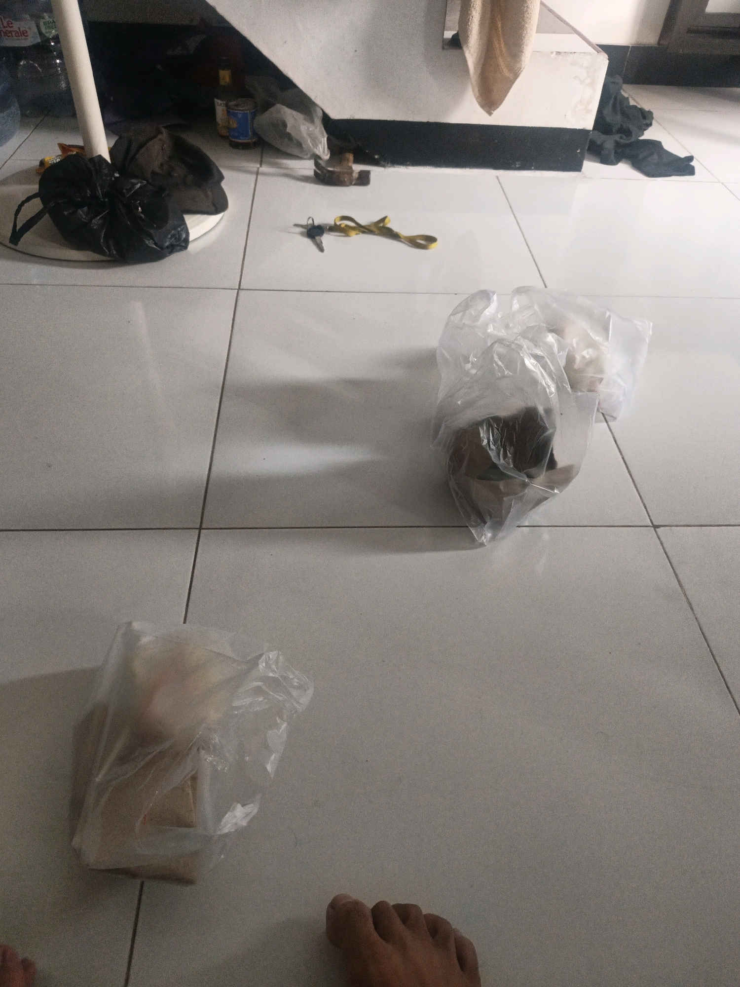 In a room with tiled flooring, likely in North Jakarta, Indonesia, several objects are scattered. In the foreground, a person's feet are visible, suggesting they are sitting or standing. To the left, a black trash bag sits on a white circular base, next to a white pole. Further back, a pair of yellow scissors and keys lie on the tiles. A clear plastic bag filled with items is positioned in the middle of the room, and another similar bag is located to the bottom left. The background reveals a dark, cluttered area with bottles, cans, and dark fabric.