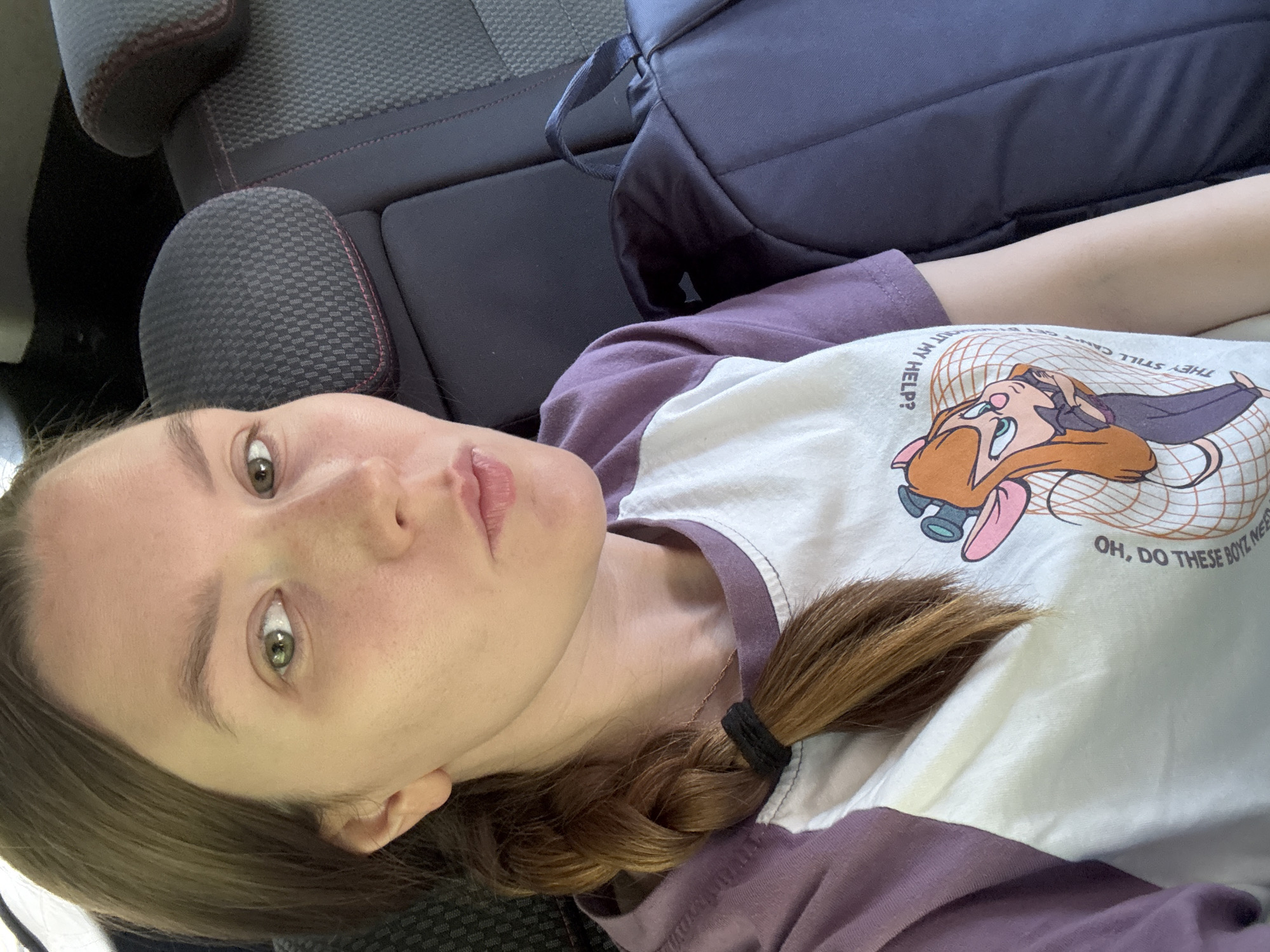 A female subject is depicted from the chest up, seated in the interior of a vehicle, likely in the backseat. Her gaze is directed forward toward the camera. She has light-colored skin, light green eyes, and medium brown hair styled in a braid over her left shoulder, secured with a black elastic tie. Her lips are slightly pursed. She is wearing a raglan-style t-shirt with a white main body and purple short sleeves. The white section of the shirt features a cartoon graphic of a female mouse character, partially obscured, and inverted text including phrases like "OH, DO THESE BOYZ NEED MY HELP?". A thin, light-colored necklace is visible around her neck. The background consists of dark grey or black car seats with a subtle patterned texture and red accent stitching on the headrests. To her right, a dark blue or black backpack rests on the seat beside her. The lighting is diffused, consistent with an indoor or shaded environment. There are no visual cues within the image to specify the geographic location as Izhevsk, Russia.