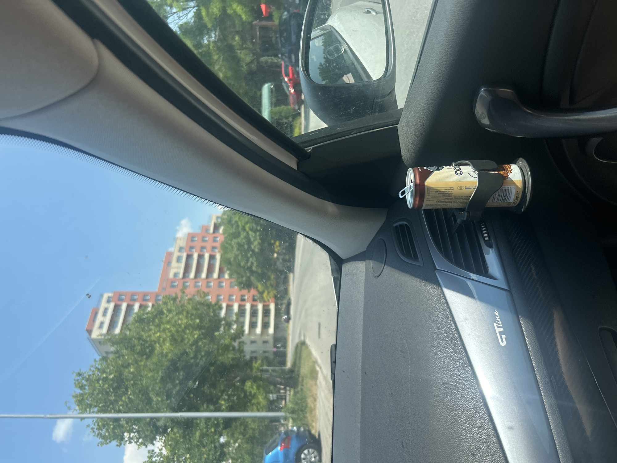 The image shows the interior of a car, looking out through the windshield and side window. A can of "COCA-COLA ZERO SUGAR" is held in a black car accessory attached to an air vent. The can has text in multiple languages, including "250MLE" and nutritional information.
Outside the car, the scene is a street with buildings and trees. A multi-story building with a distinctive architectural style, likely residential, is visible in the background. The sky is clear and blue, suggesting a sunny day. The time of day appears to be daytime, with bright sunlight casting shadows.
A side mirror is visible on the right side of the frame, reflecting a portion of the car's interior and the outside environment. A dark gray car door handle is also prominent in the foreground. The car's dashboard is visible, with a textured surface and a "GT Line" emblem.
The overall impression is a snapshot of a moment during a car journey on a bright, clear day in an urban setting, possibly Plovdiv, Bulgaria, given the location context provided. There are no people visible in the image. The activity is simply the state of the car interior and the view outside.