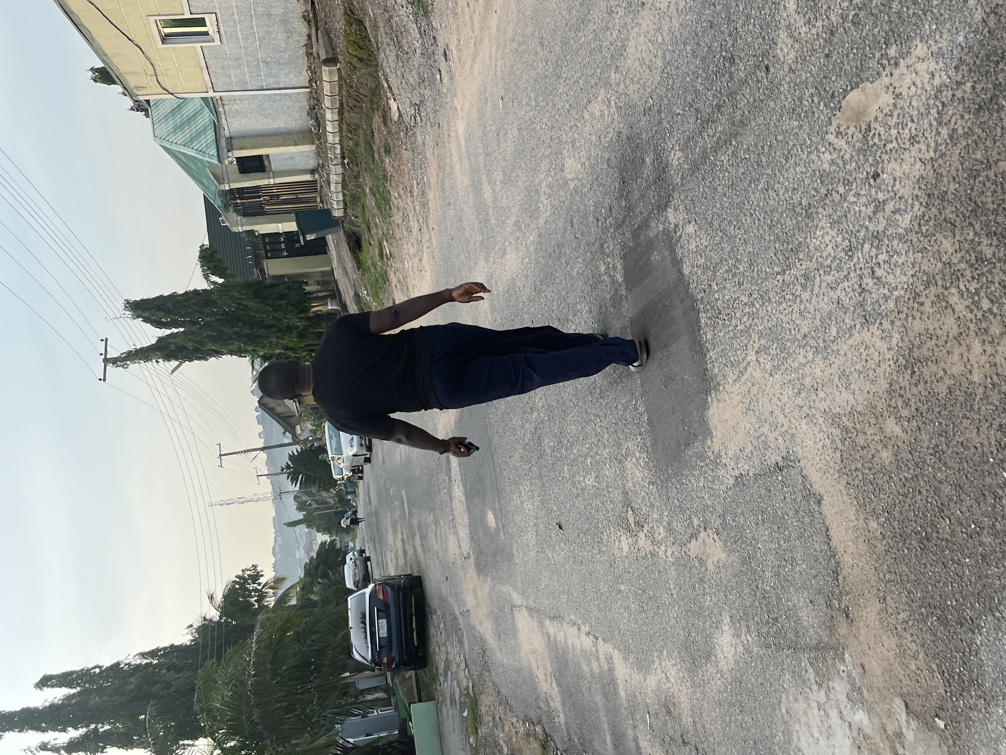 An outdoor, eye-level, full shot captures a man from behind walking away from the camera down a narrow, paved road. The man is wearing a black t-shirt and dark blue pants. He is holding a black smartphone in his right hand. The road has patches of lighter colored material and visible cracks, with a slight incline. On the left side of the road, several cars are parked, including a dark sedan. Beyond the cars, palm trees and tall evergreen trees line the street. On the right side of the road are several residential buildings with light-colored exteriors, some with green roofs, and fences. The sky is overcast and light blue. In the distance, hills can be seen. The scene appears to be in a residential area of Abuja, Nigeria, given the architectural style of the buildings and the presence of palm trees.