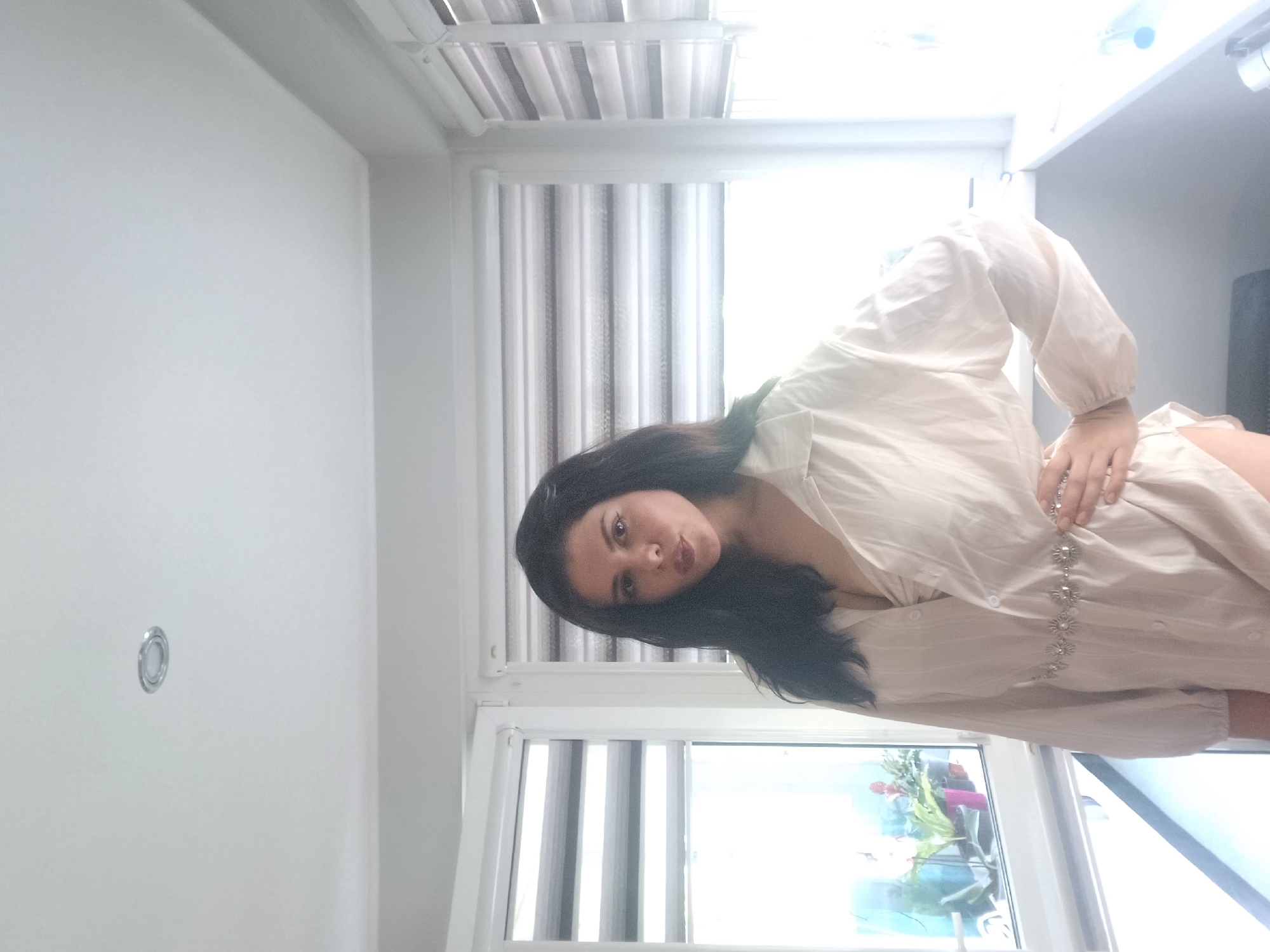 A woman with long, dark hair is posing indoors in front of a window with closed blinds. She is wearing a long-sleeved, light-colored dress. The room has white walls and a ceiling with a light fixture. The exact location in Sofia, Bulgaria is impossible to determine without more information.