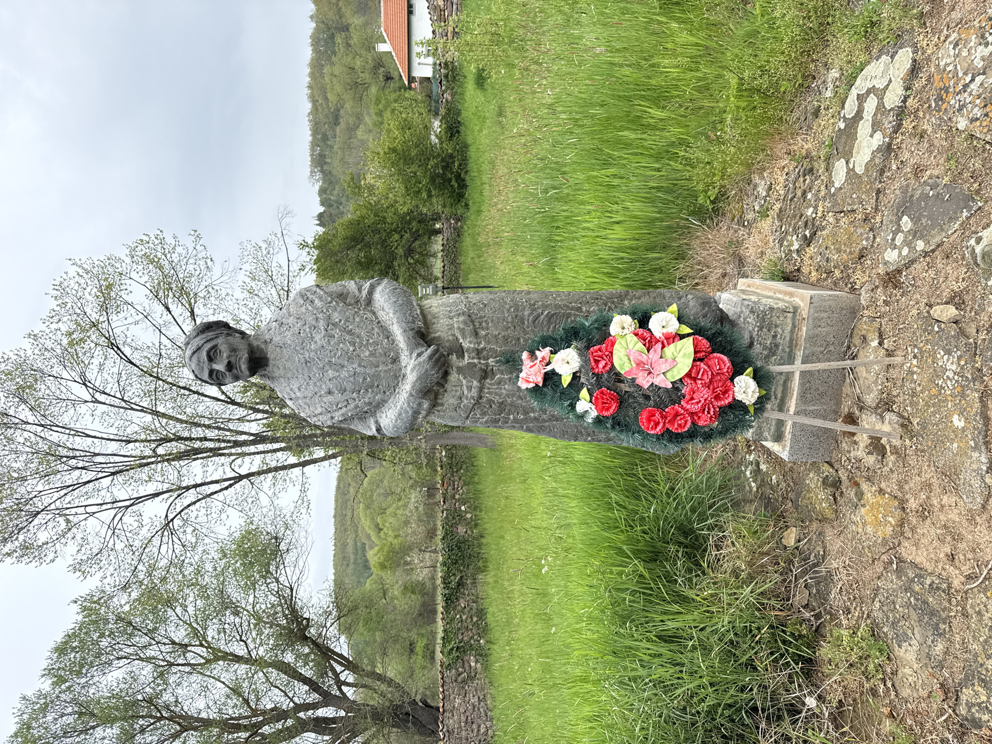 The image shows a stone statue of a woman, possibly a religious figure, located outdoors. A colorful floral wreath adorns the base of the statue. The setting appears to be a grassy field with a stone wall, trees, and a glimpse of a building in the background. The statue is presumably located in Kovachevtsi, Bulgaria.