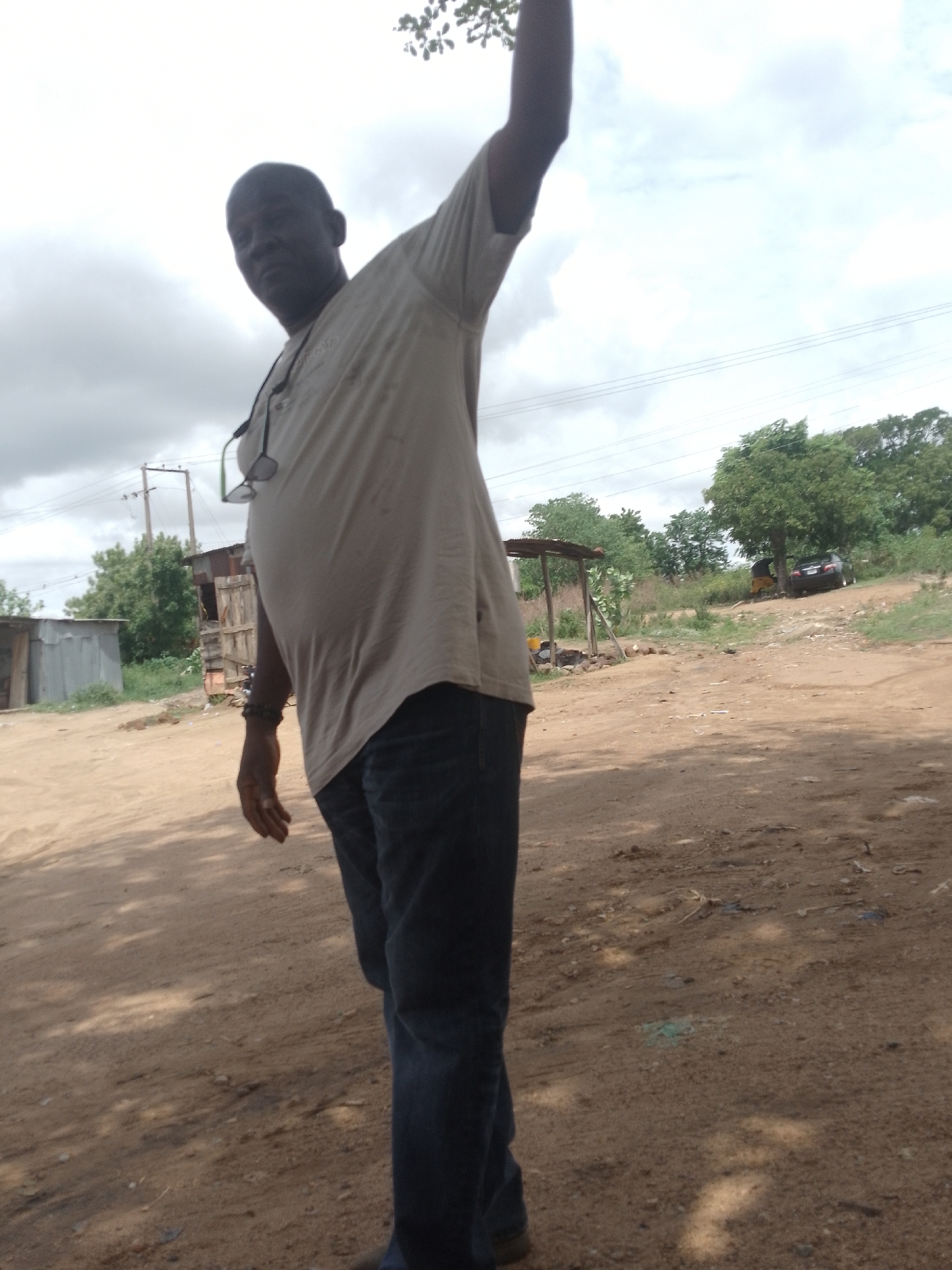 An adult male stands in a dirt lot, arm raised. He wears jeans and a light-colored t-shirt. A pair of eyeglasses hangs on his shirt. In the background, there are rudimentary structures, trees, and an overcast sky. The location is Adankolo, Nigeria. A vehicle is also visible in the distance.