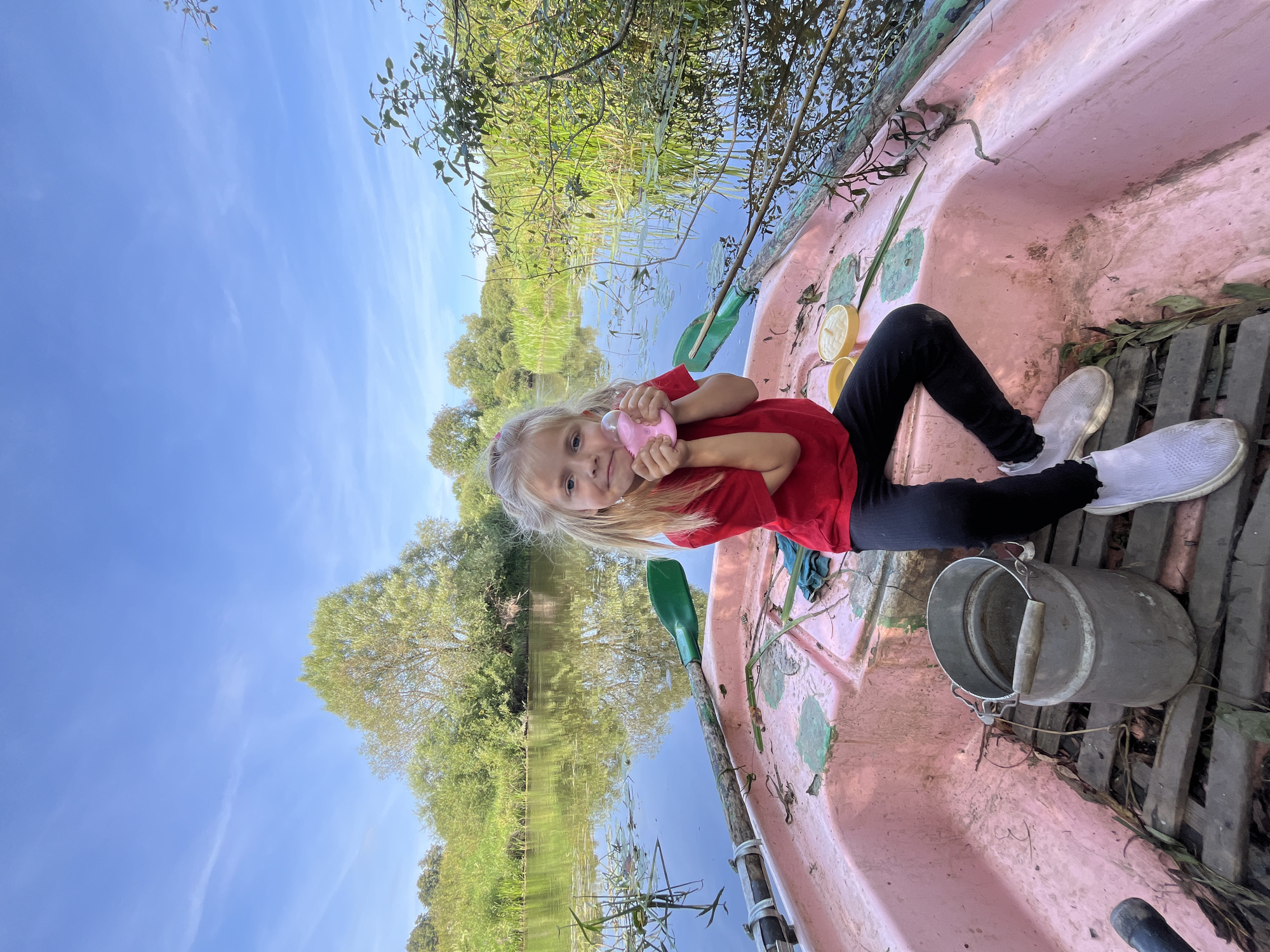 A young girl with blonde pigtails sits in a pink rowboat on a calm river. She is wearing a red t-shirt and black leggings. In her hands, she holds a small pink toy. The boat has green oars, a metal bucket, and a wooden floor. Lush green trees and reeds line the riverbanks. The sky is blue with a few wispy clouds. The scene appears to be in a rural, natural setting, possibly in or near the city of Sharkawshchyna, Belarus.