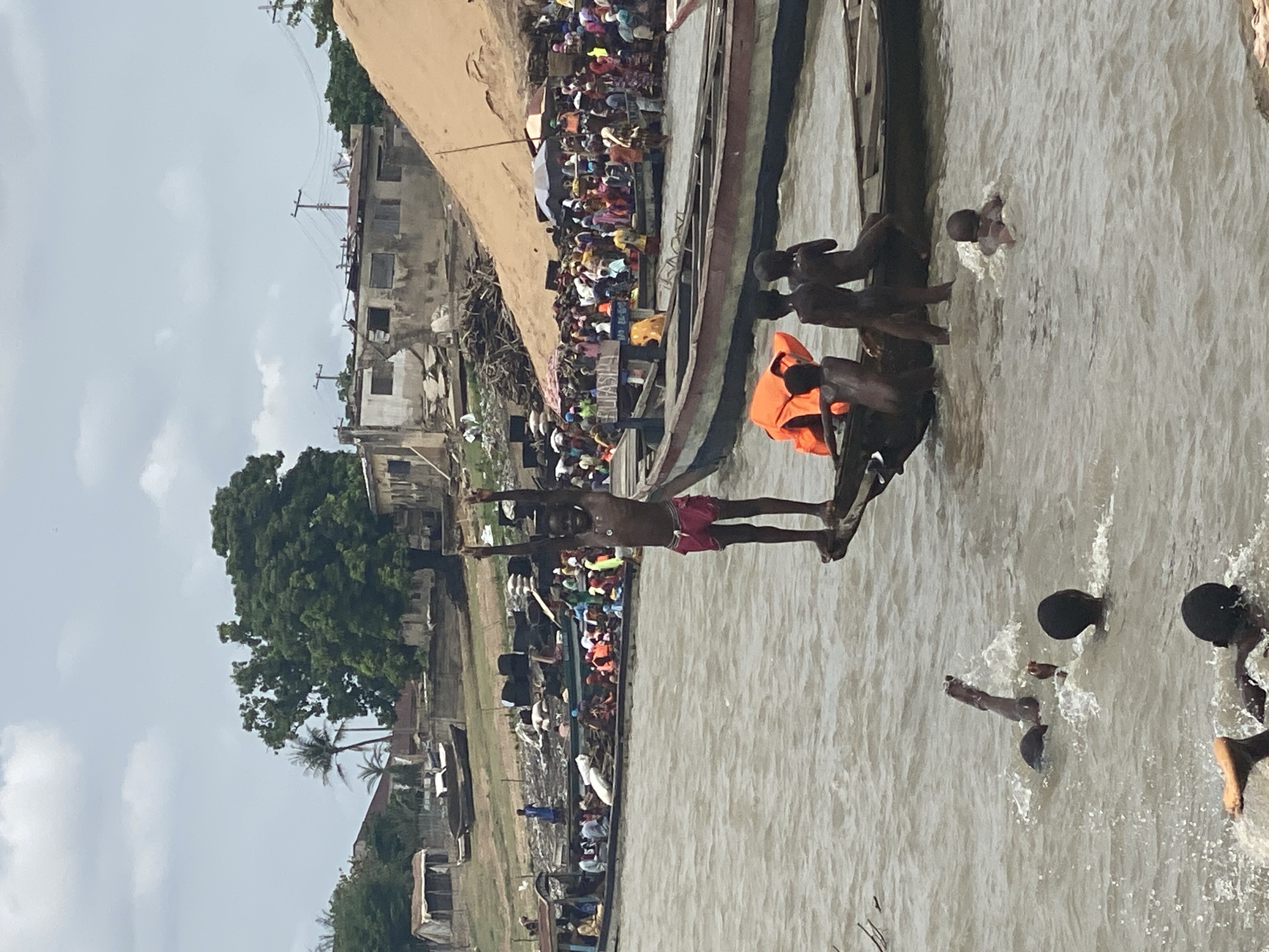 The image depicts a scene on the Niger River in Lokoja, Nigeria. The river is turbid and filled with swimmers, likely children, their heads visible as they swim and splash in the water. Along the shoreline, crowds of people gather on the riverbank and on boats. The boats, long and narrow, are crowded with spectators. The shoreline is lined with structures ranging from dilapidated buildings to trees. In the foreground, a young man stands poised to dive into the water from the edge of a boat. Several other people are also on the boat, some wearing orange life jackets. The overall scene suggests a crowded event or festival taking place on the river.